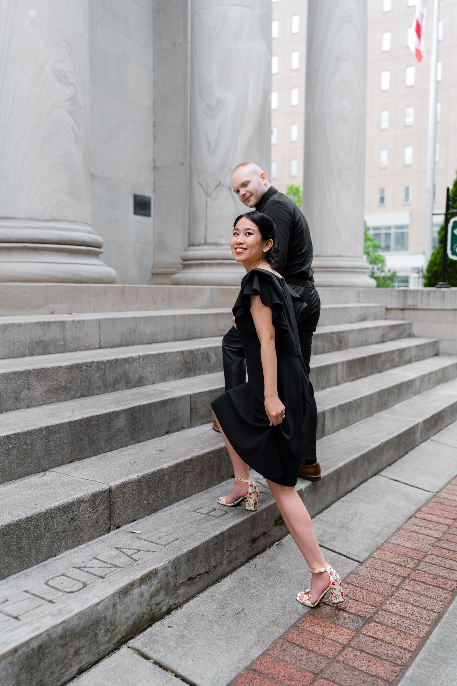 Couple in black outfits posing on stone steps of a grand building with large columns.