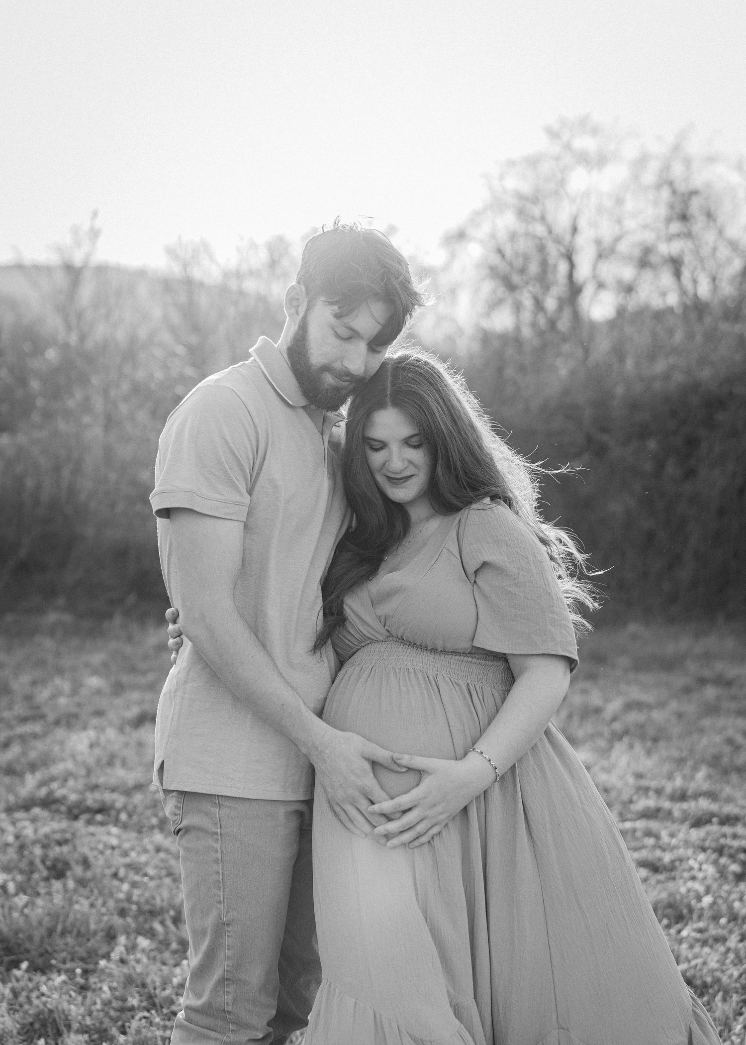 A black and white maternity photo of a couple embracing outdoors, both looking down at her baby bump.