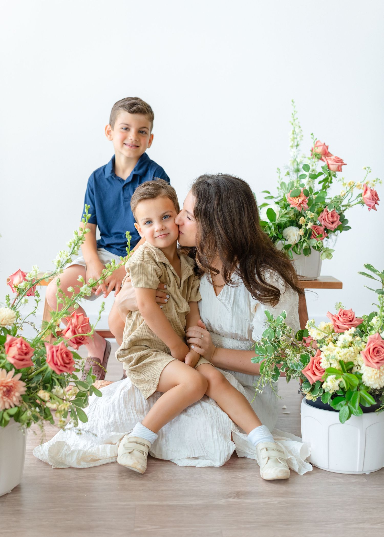 A mother kisses her young son while another boy smiles among pink roses in a bright studio setting.