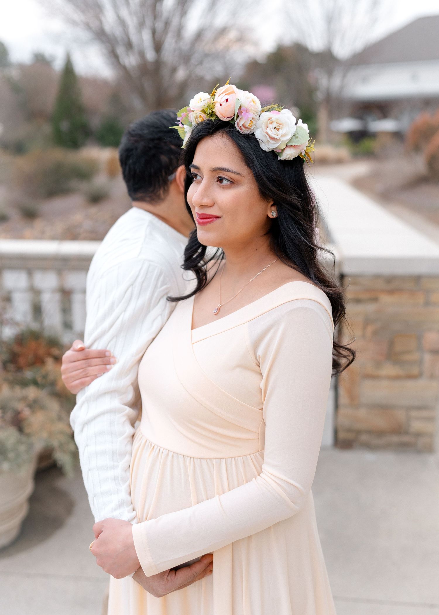 Pregnant woman in cream gown and floral crown embraced by partner outdoors.