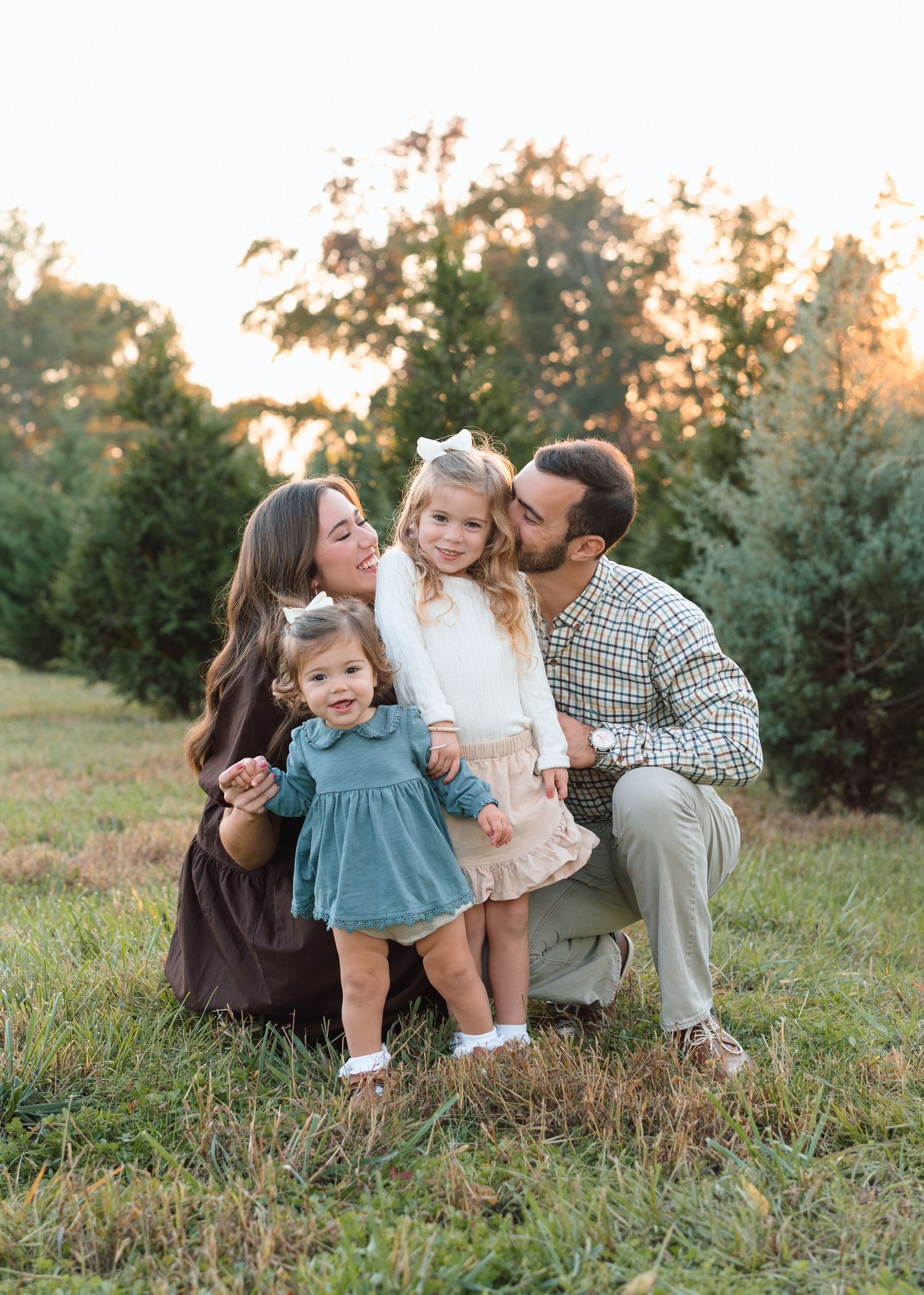 Loving family of four sharing kisses with two young daughters at golden hour near evergreen trees.