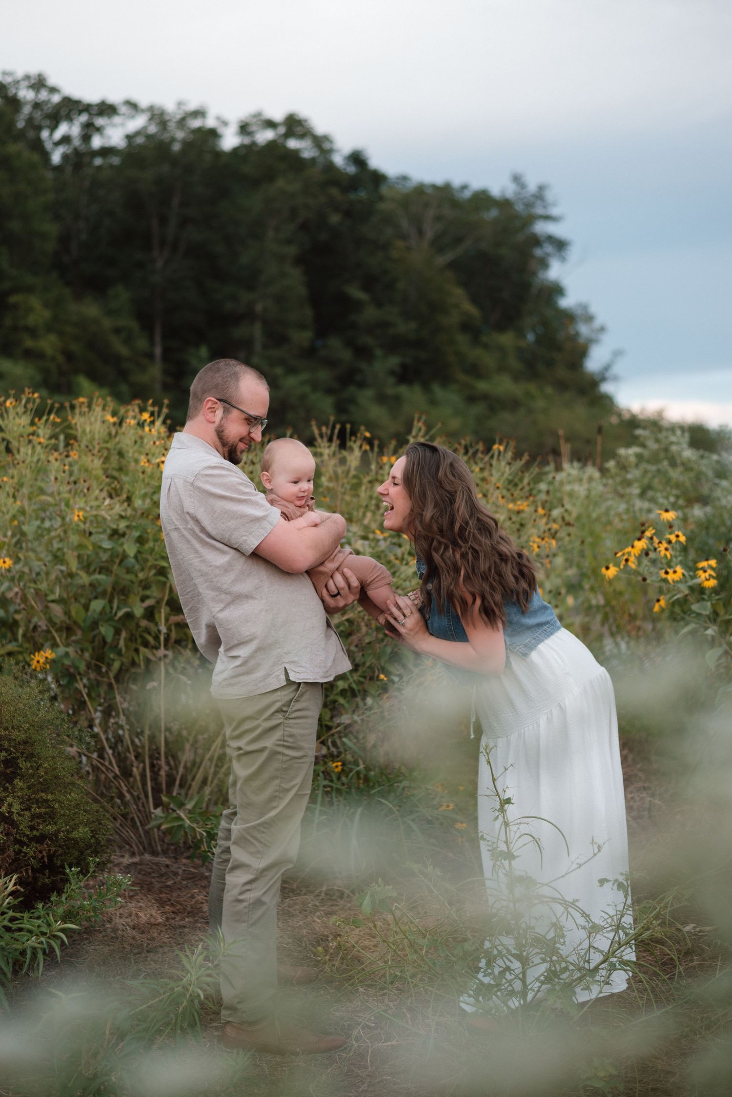 Happy family with baby boy surrounded by wildflowers and yellow blooms in a lush green garden setting.