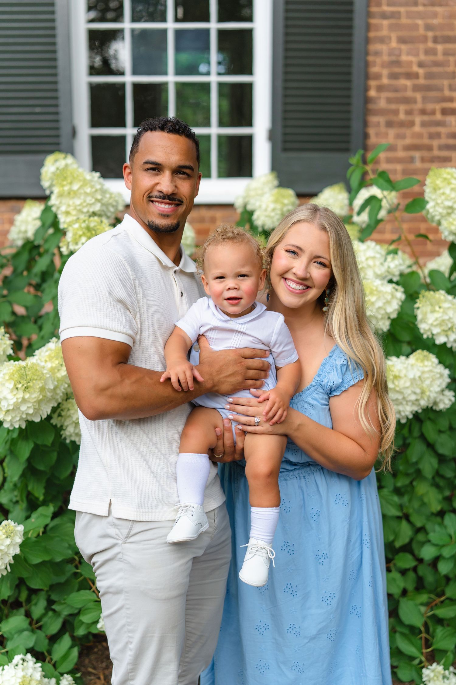 Happy interracial family posing outdoors with baby boy in front of white hydrangeas and brick home.