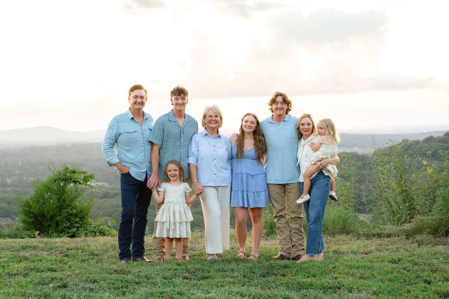 A joyful multigenerational family poses together on a green hillside at sunset with scenic landscape views behind them.