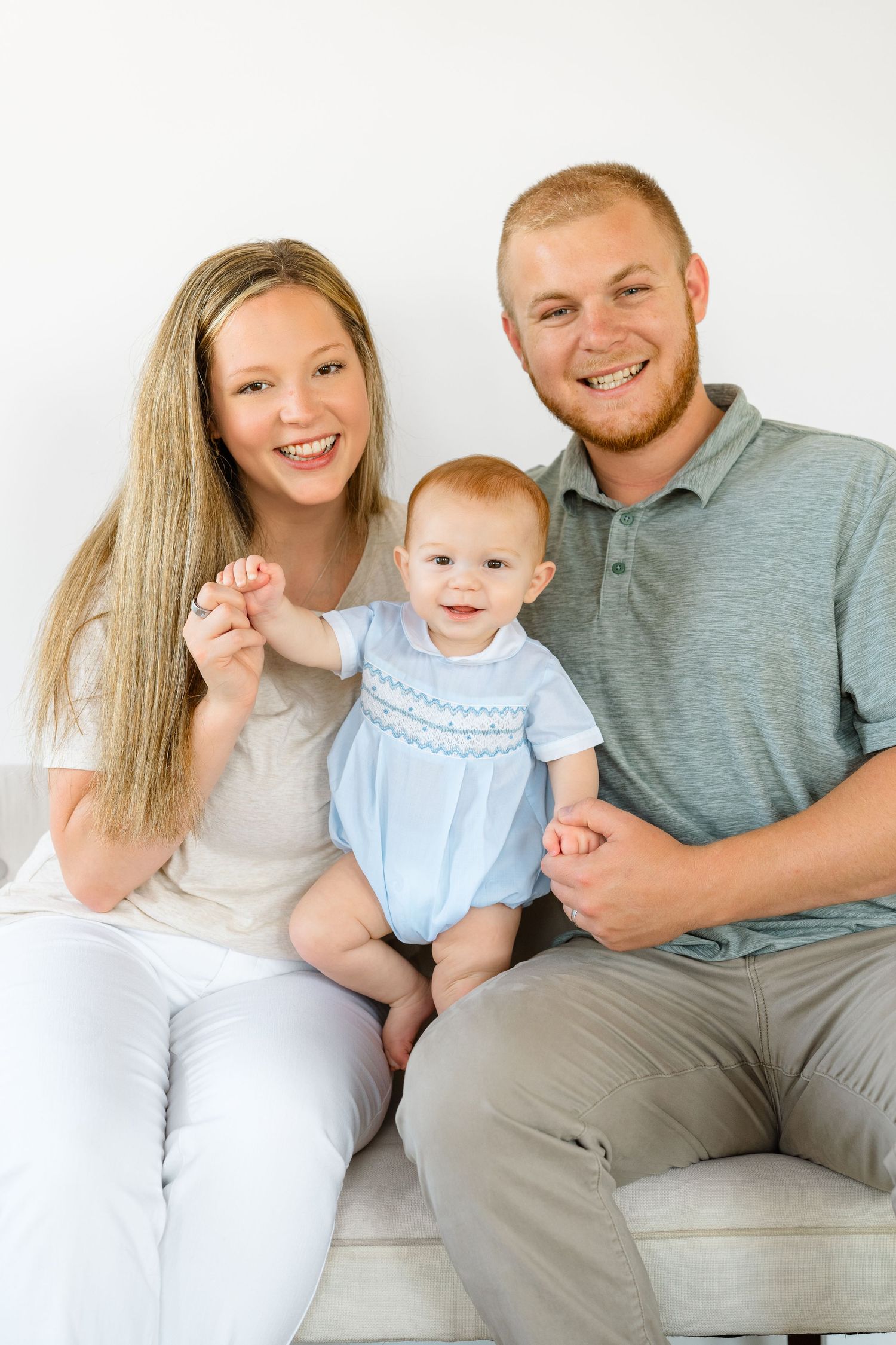 Smiling young family with redhead baby in blue romper sitting on white couch.