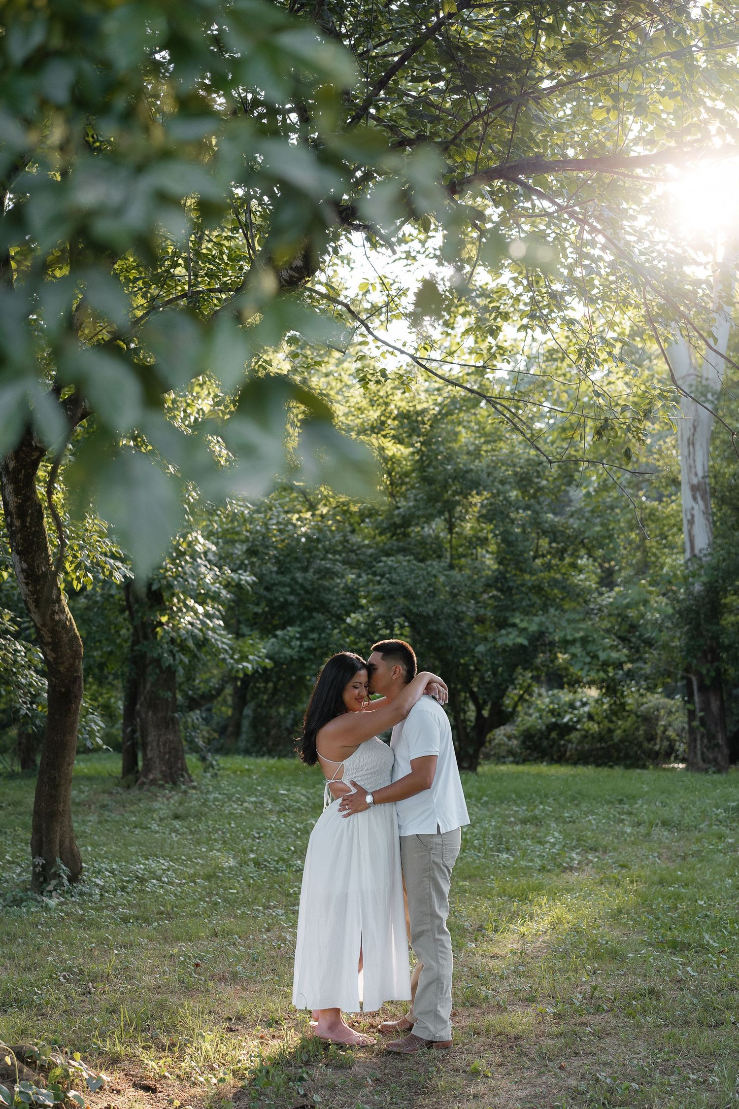 Couple embracing in a sunlit green park, woman in white dress, romantic outdoor photo session.