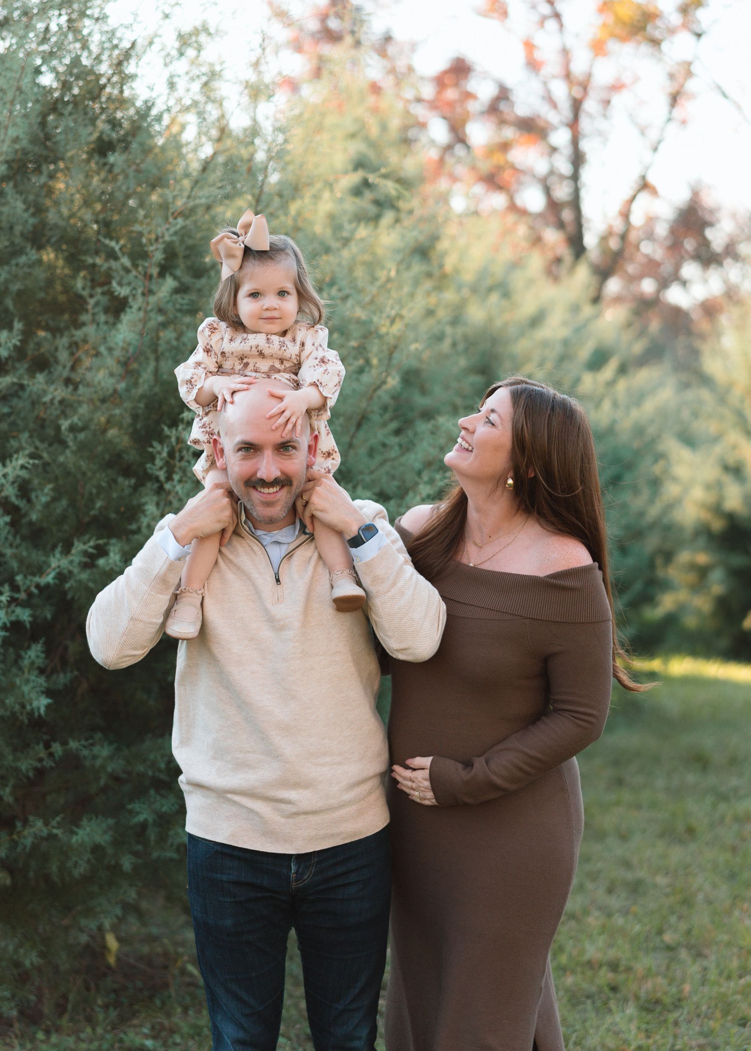 Father with toddler on shoulders and pregnant wife laughing outdoors among evergreen trees in fall.