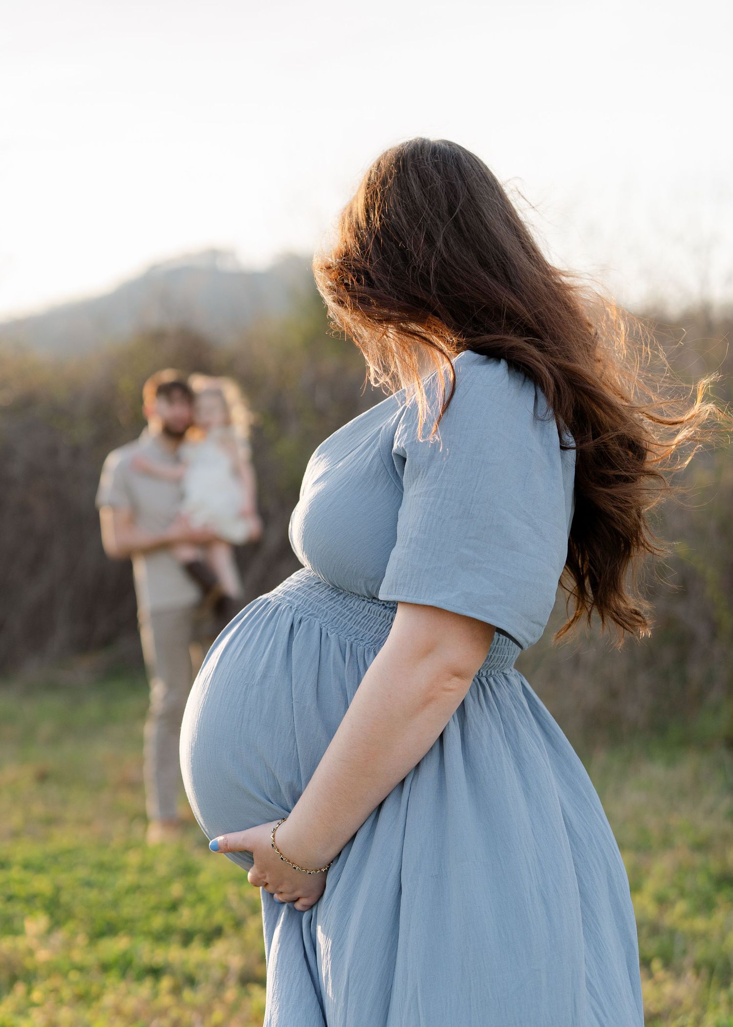 A pregnant woman in a blue dress stands in a field while her partner holds their child in the background.