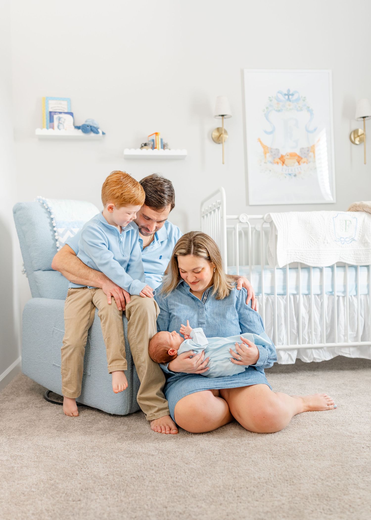Family with newborn baby and toddler son in blue nursery with glider chair and white crib.