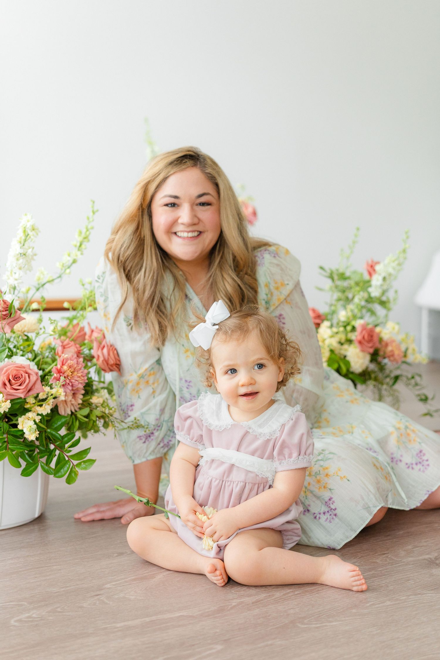 A mother in a floral dress smiles with her toddler daughter sitting on a floor among pink flowers.