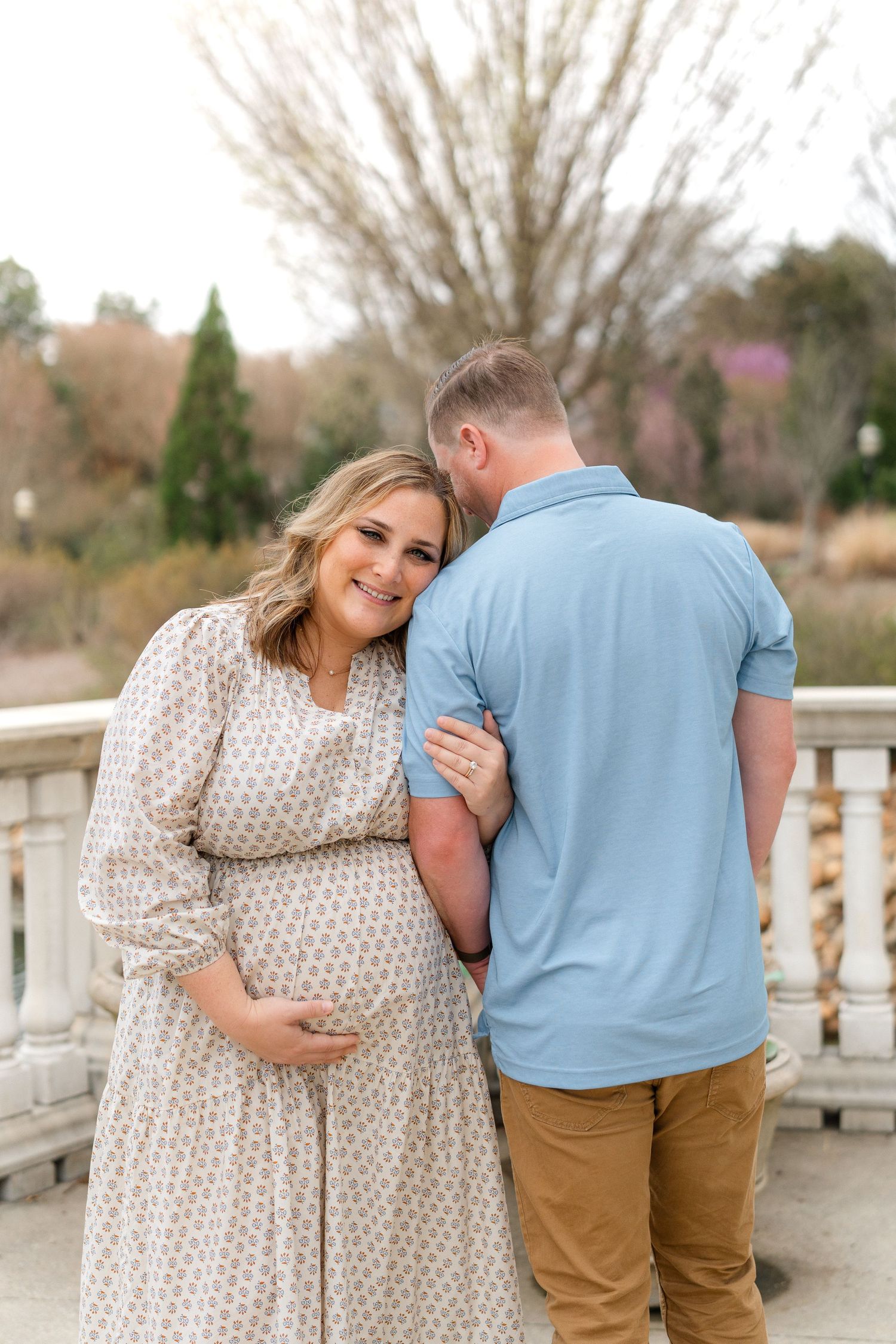 A smiling pregnant woman in a patterned dress holds her baby bump while her partner leans in close outdoors.