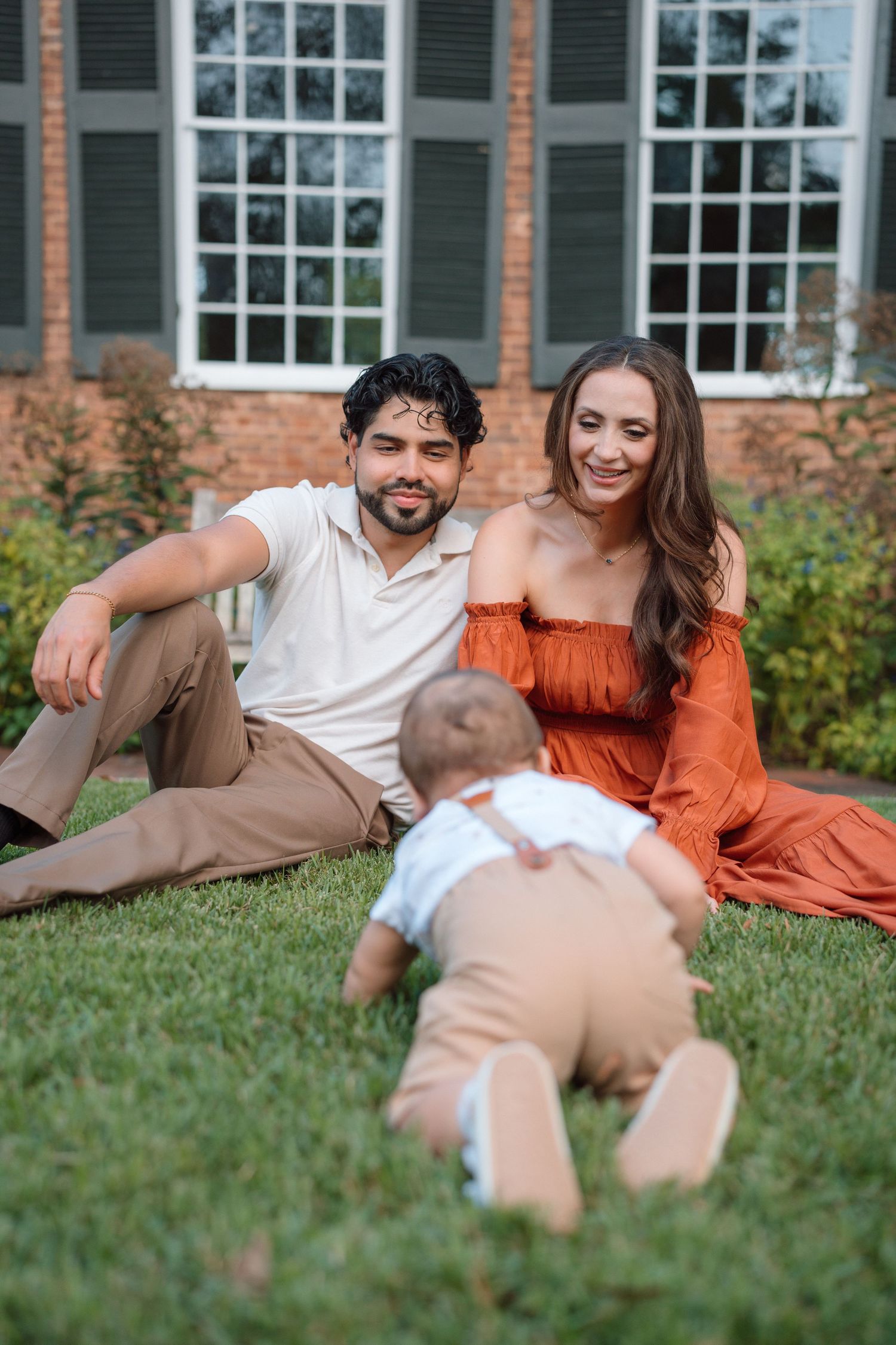 A young couple sits on green grass smiling at their crawling baby in front of a brick house.