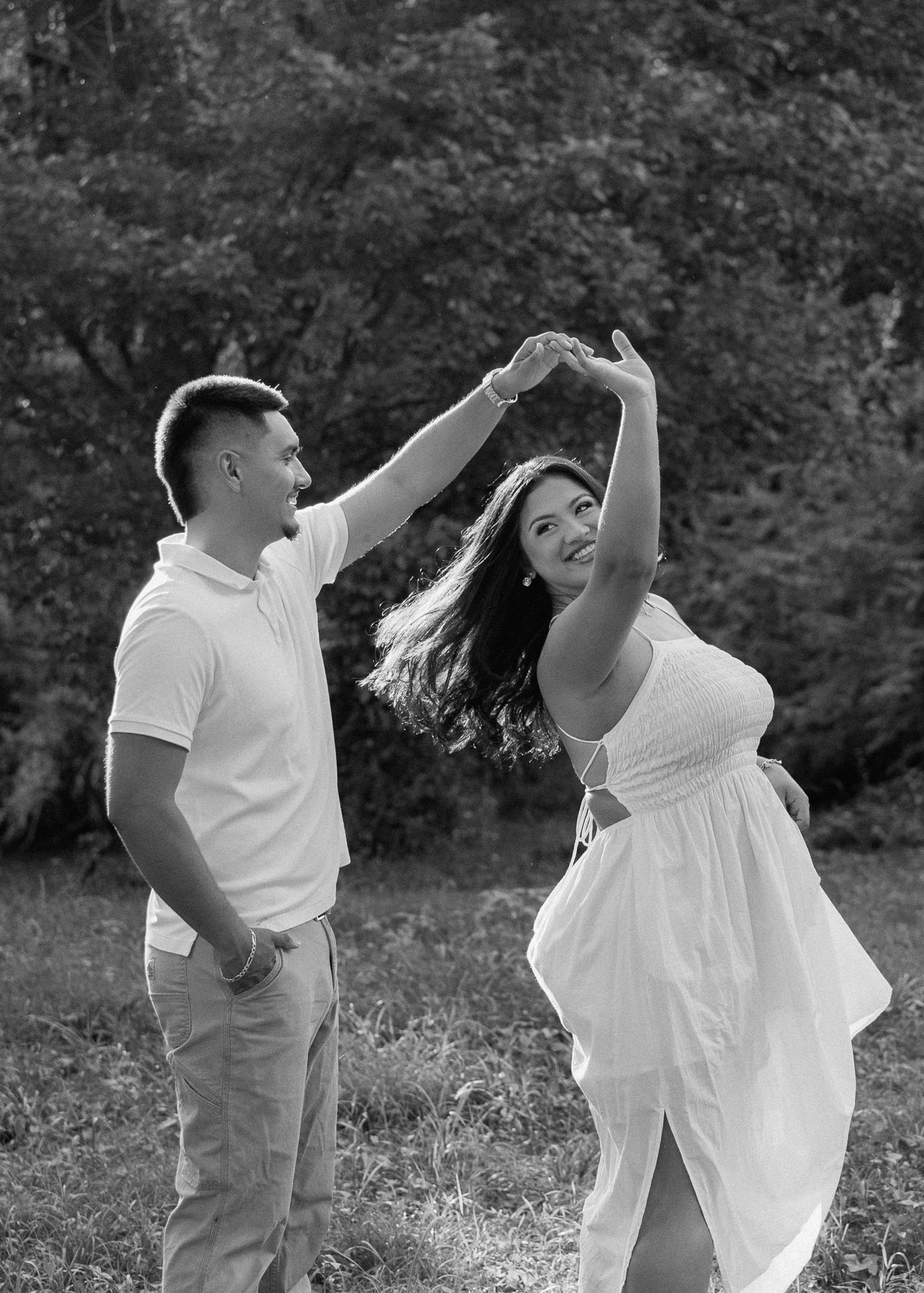 Black and white photo of a couple dancing outdoors, man spinning woman in white dress, trees in background.