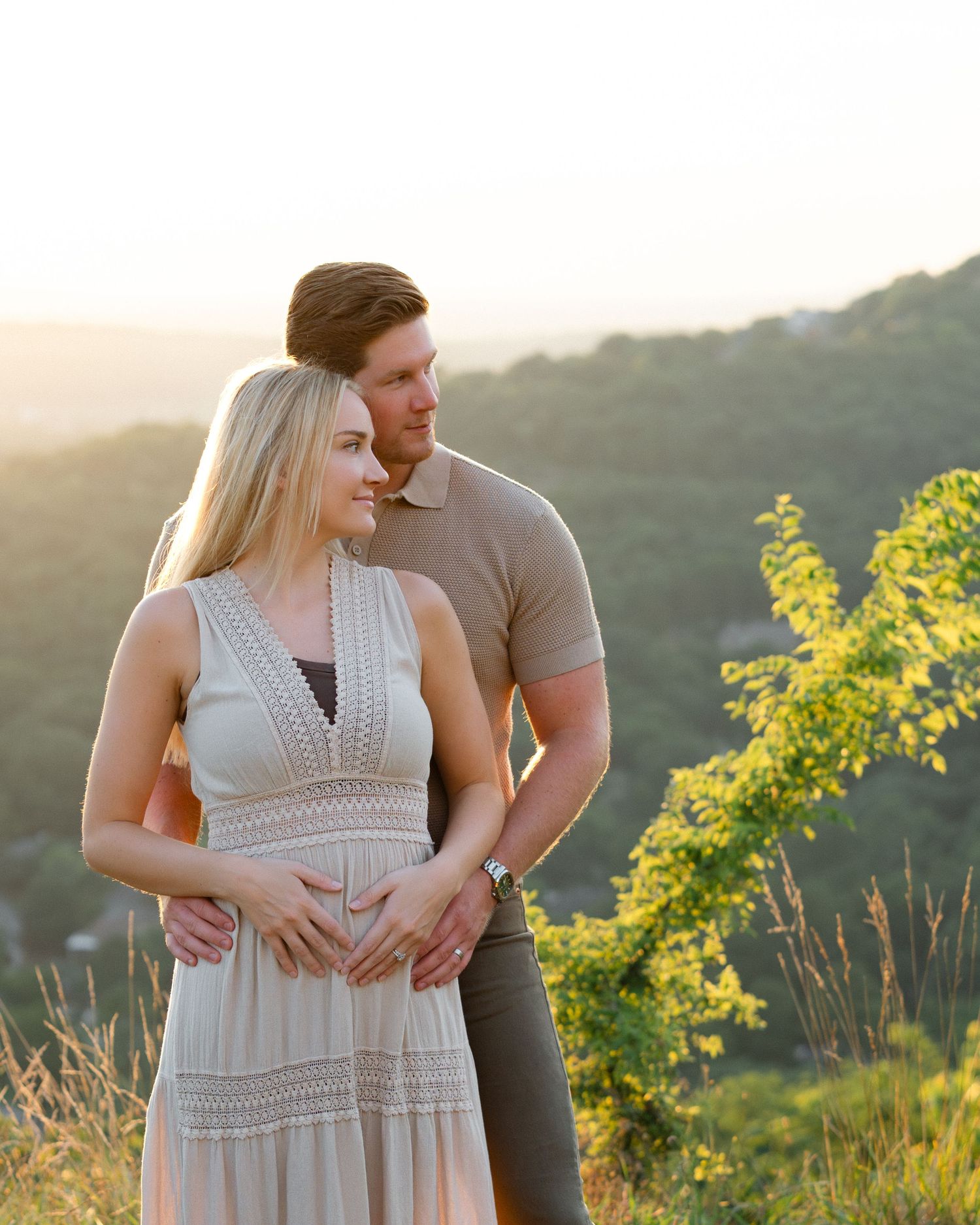 Couple poses at golden hour on a hilltop, woman in flowy dress with hands on baby bump, lush green valley behind them.