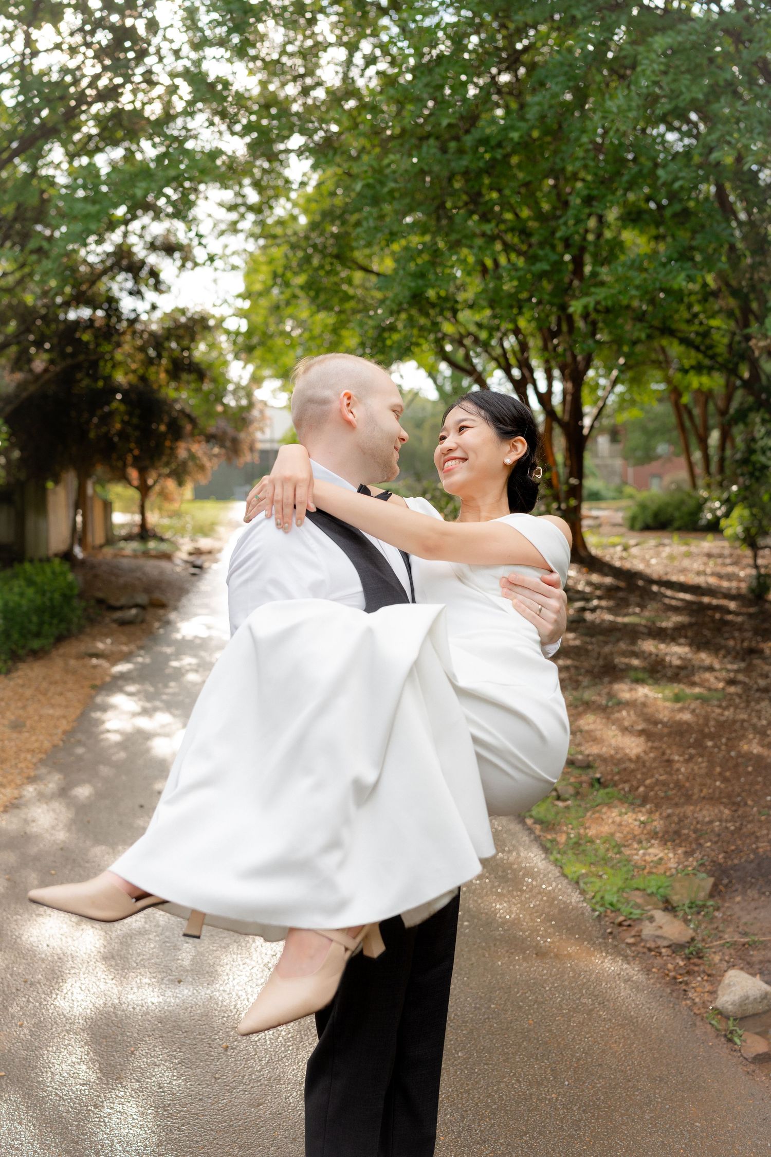 Groom carries smiling bride in white wedding dress outdoors on a tree-lined path on their wedding day.