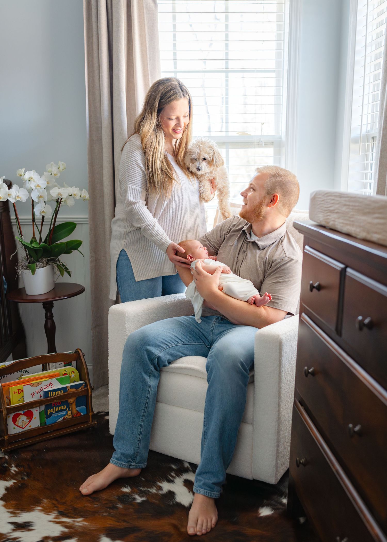 Father sits in nursery chair holding newborn while mother stands nearby holding small fluffy dog.