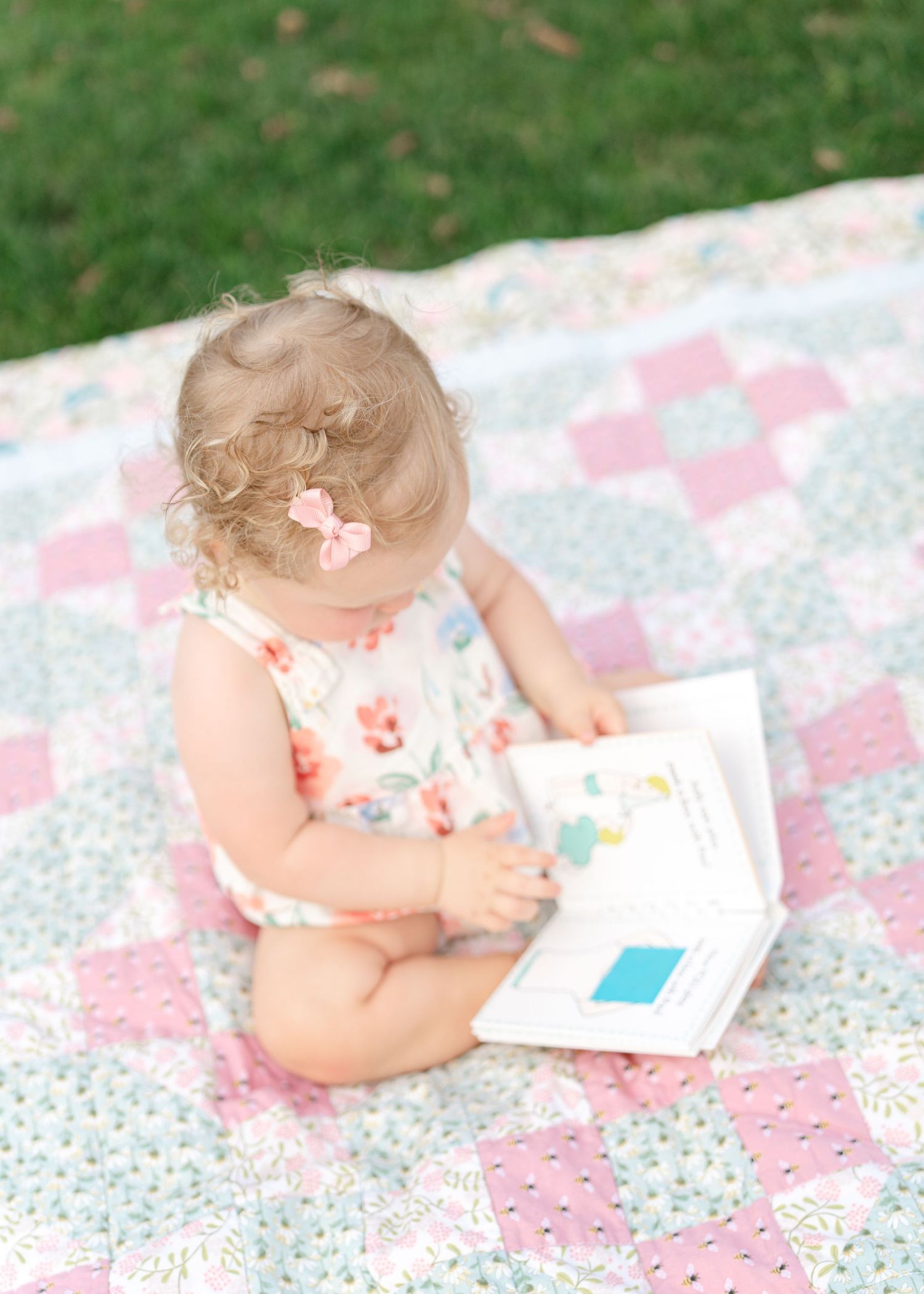 Toddler girl in floral romper reading a book on a pastel patchwork quilt outdoors.