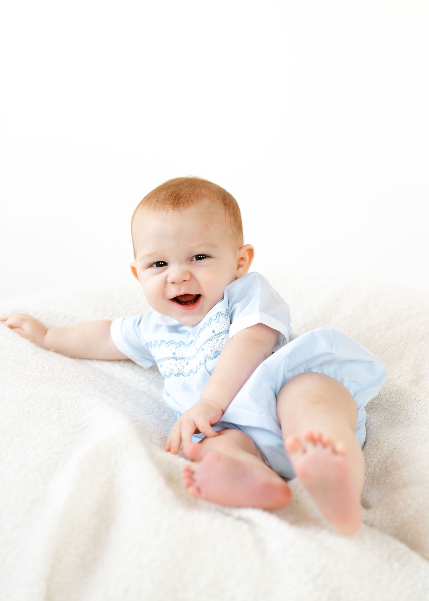 Happy baby in blue romper sitting and laughing on a soft white blanket.