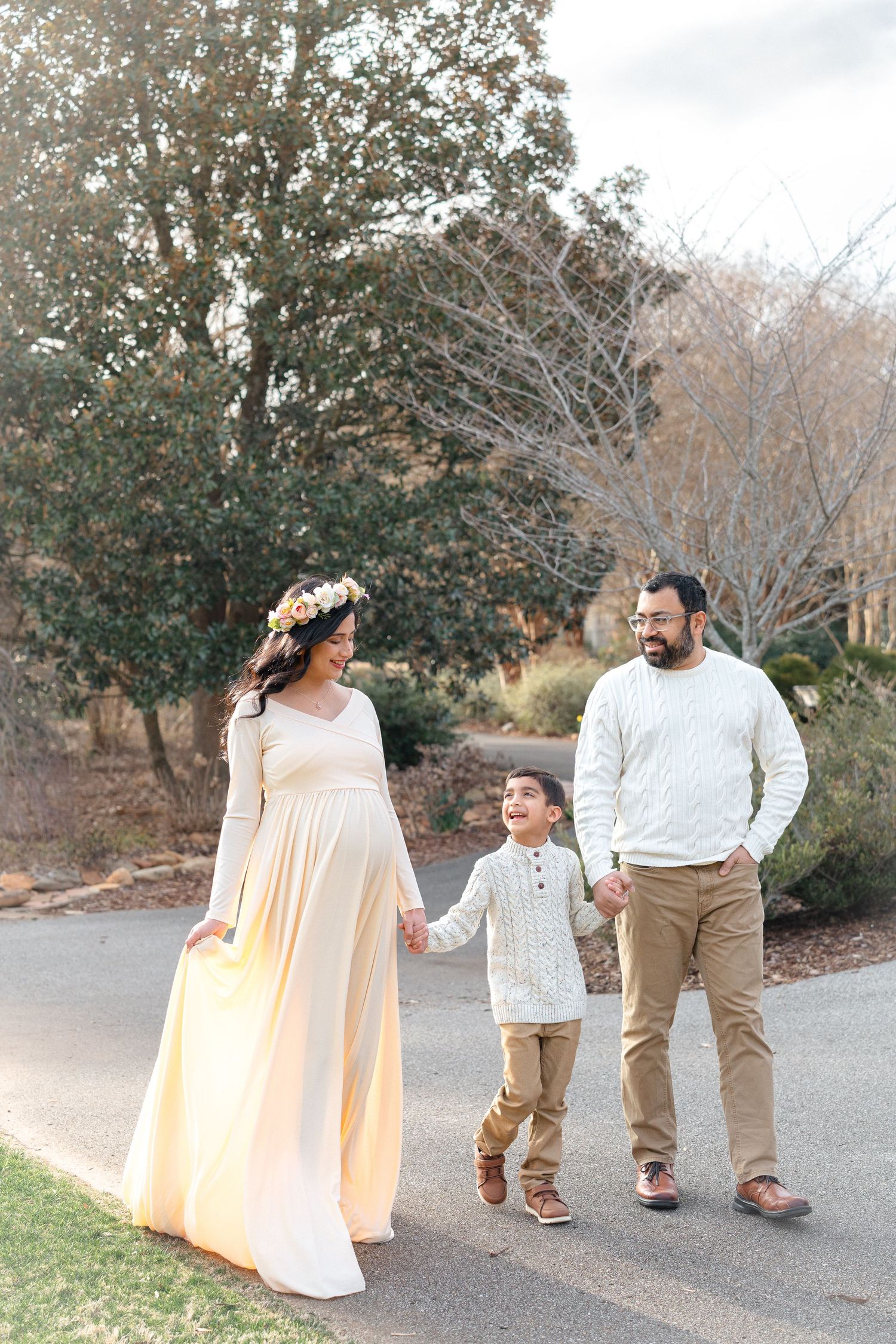 Pregnant woman in cream gown and floral crown walks with husband and son on path.