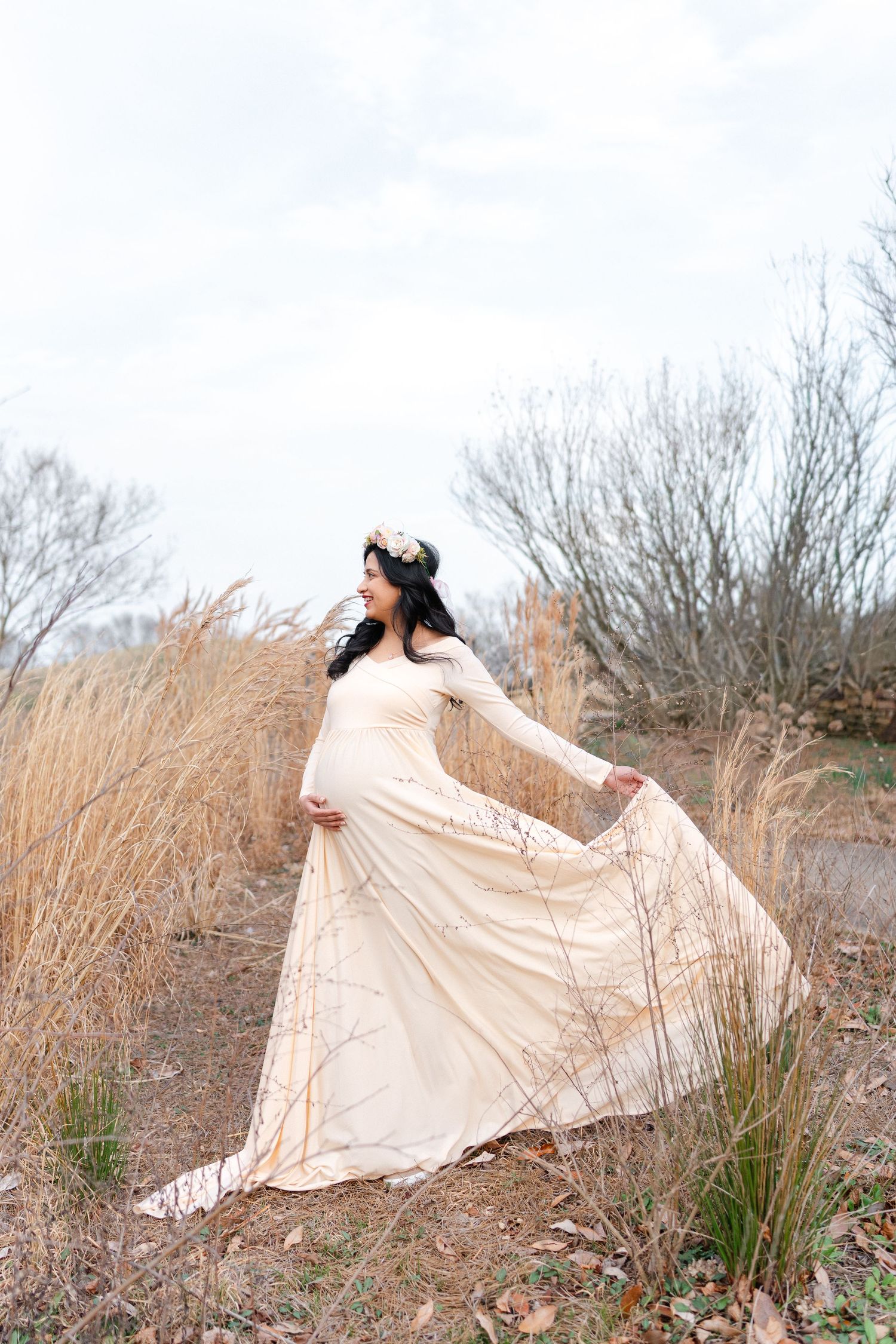 Pregnant woman in cream maternity dress and floral crown twirls in field of tall grass.