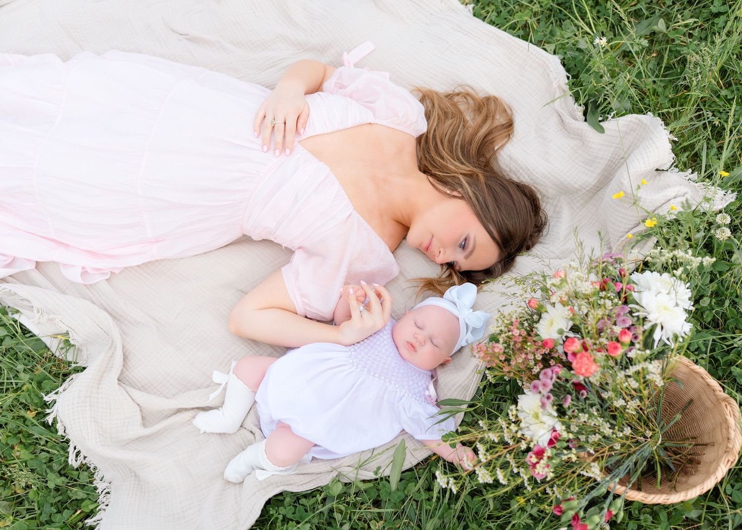 A mother in a pink dress lies on a blanket in the grass next to her newborn baby and wildflowers.