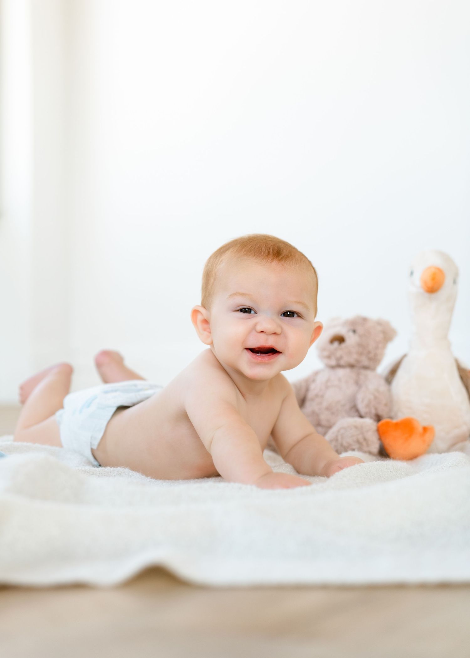 Smiling baby in diaper doing tummy time on white blanket with stuffed animal toys.
