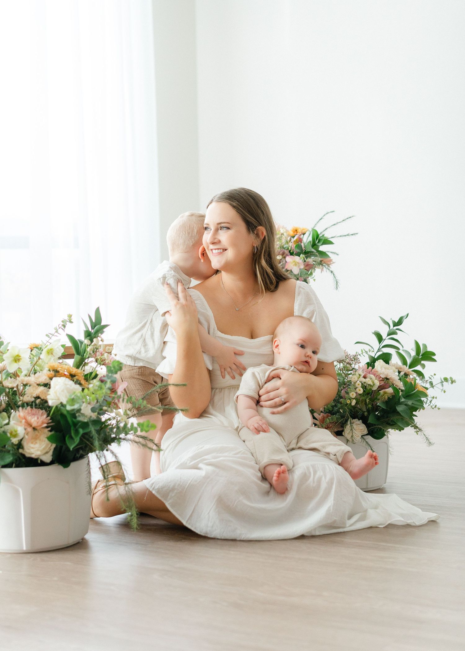 Brunette mother in white dress sits on floor smiling while holding two babies with flowers nearby.
