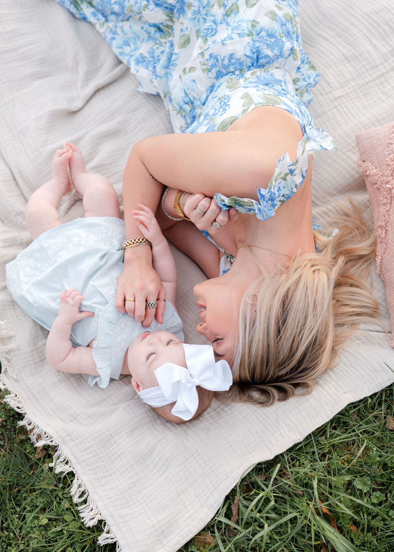 Blonde mother in blue floral dress lies beside baby girl with white bow on blanket.