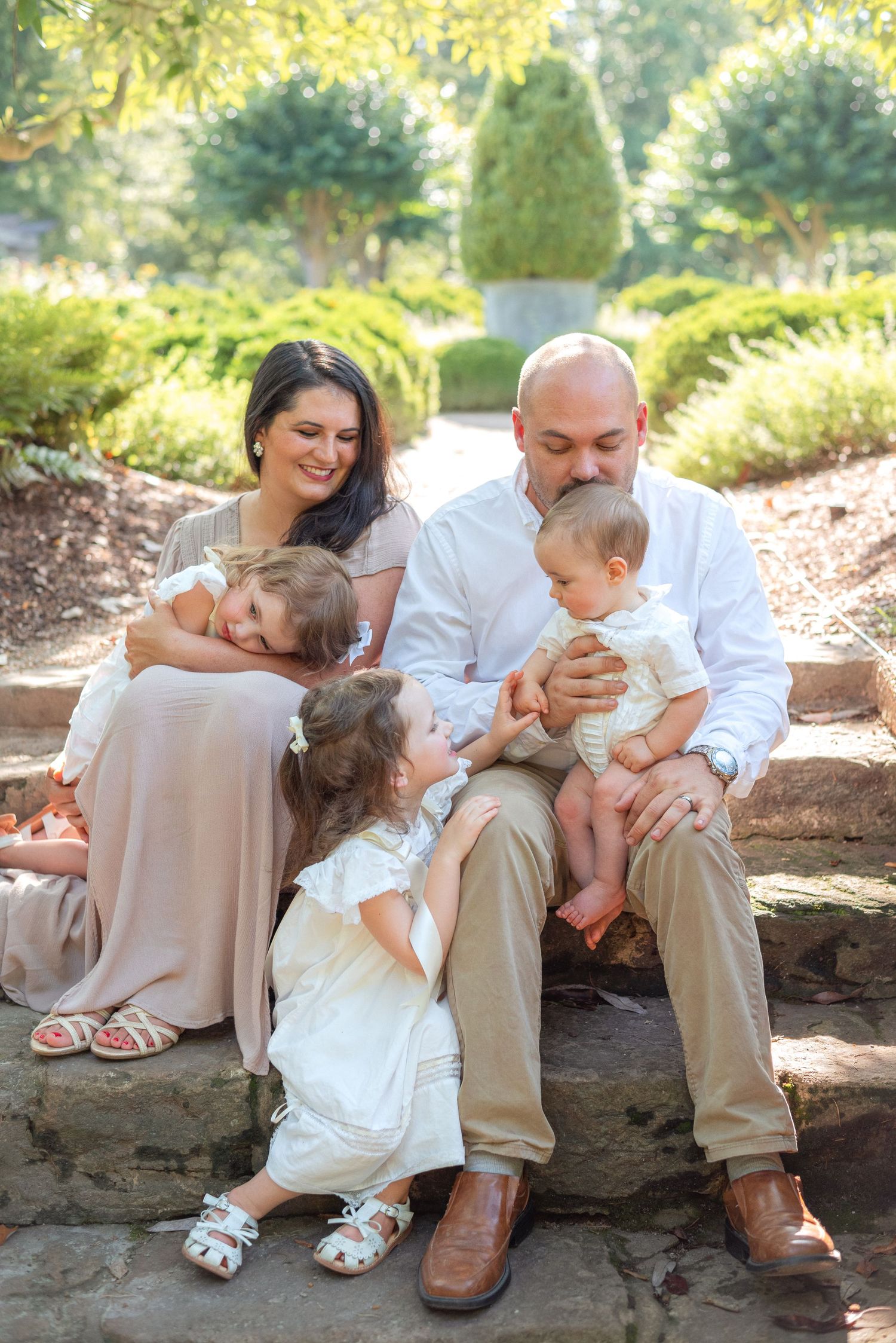 Family of five sitting on stone steps in a lush garden, parents holding children while a toddler stands between them.