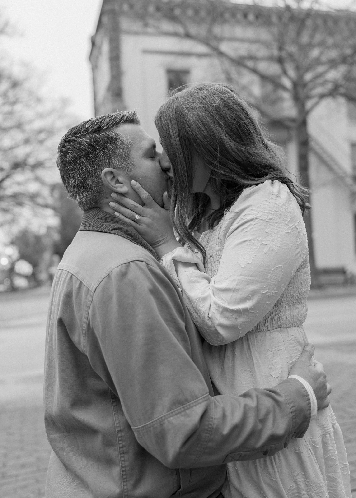 Black and white romantic photo of engaged couple sharing a kiss outdoors near a historic building.