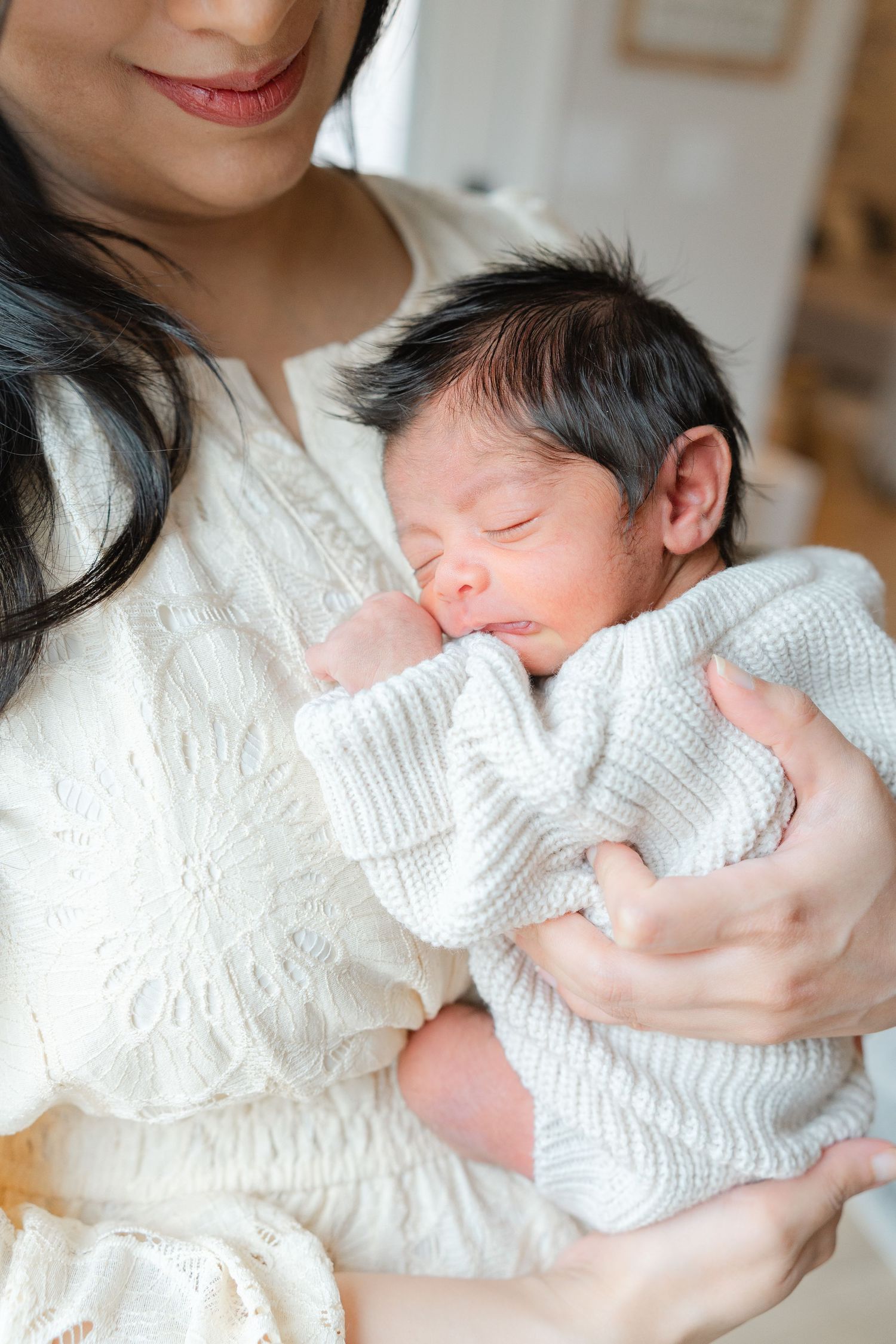 Mother in white dress tenderly holds sleeping newborn baby dressed in white knit outfit.