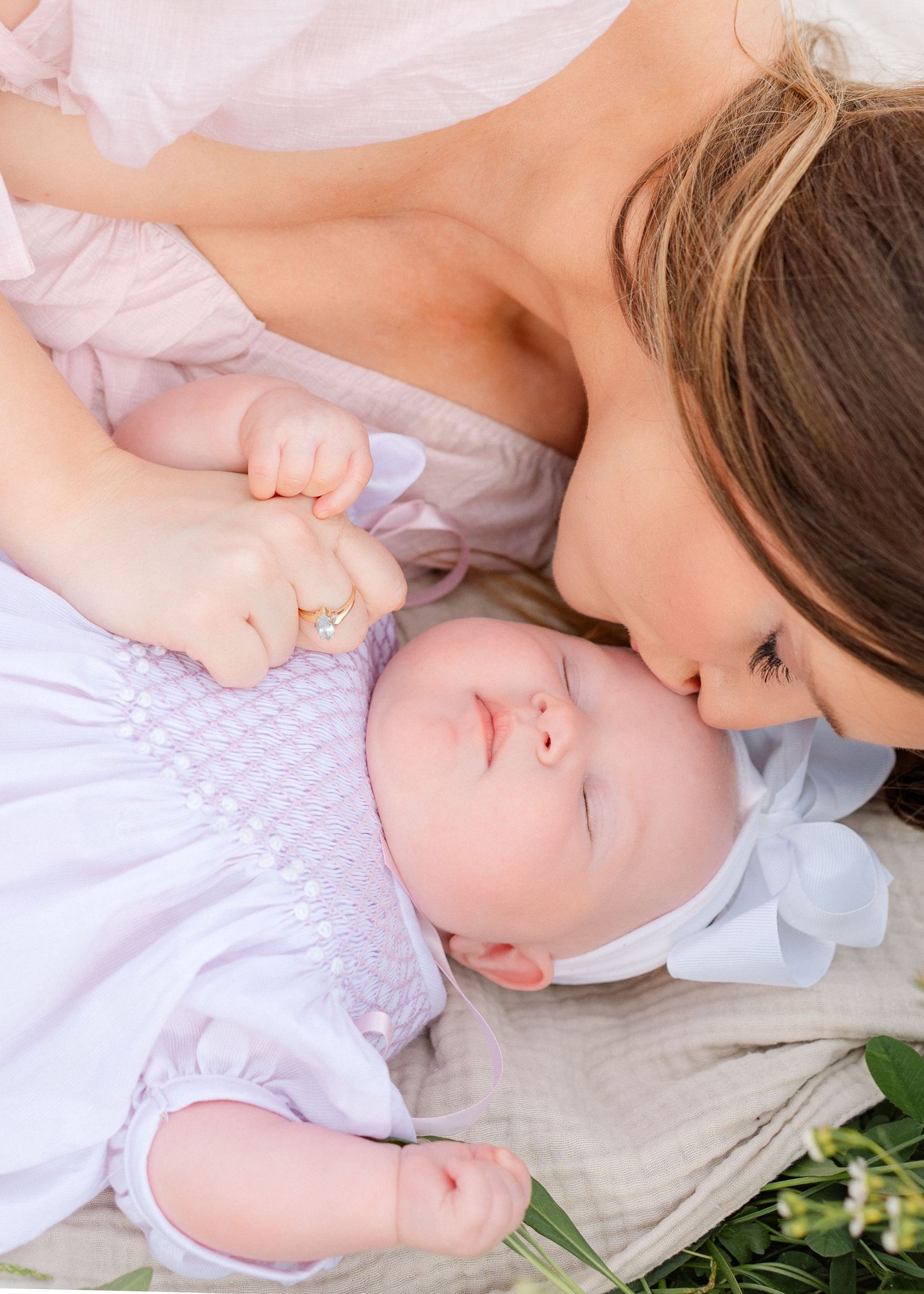 A mother tenderly kisses her smiling baby dressed in a lavender smocked outfit on a blanket.