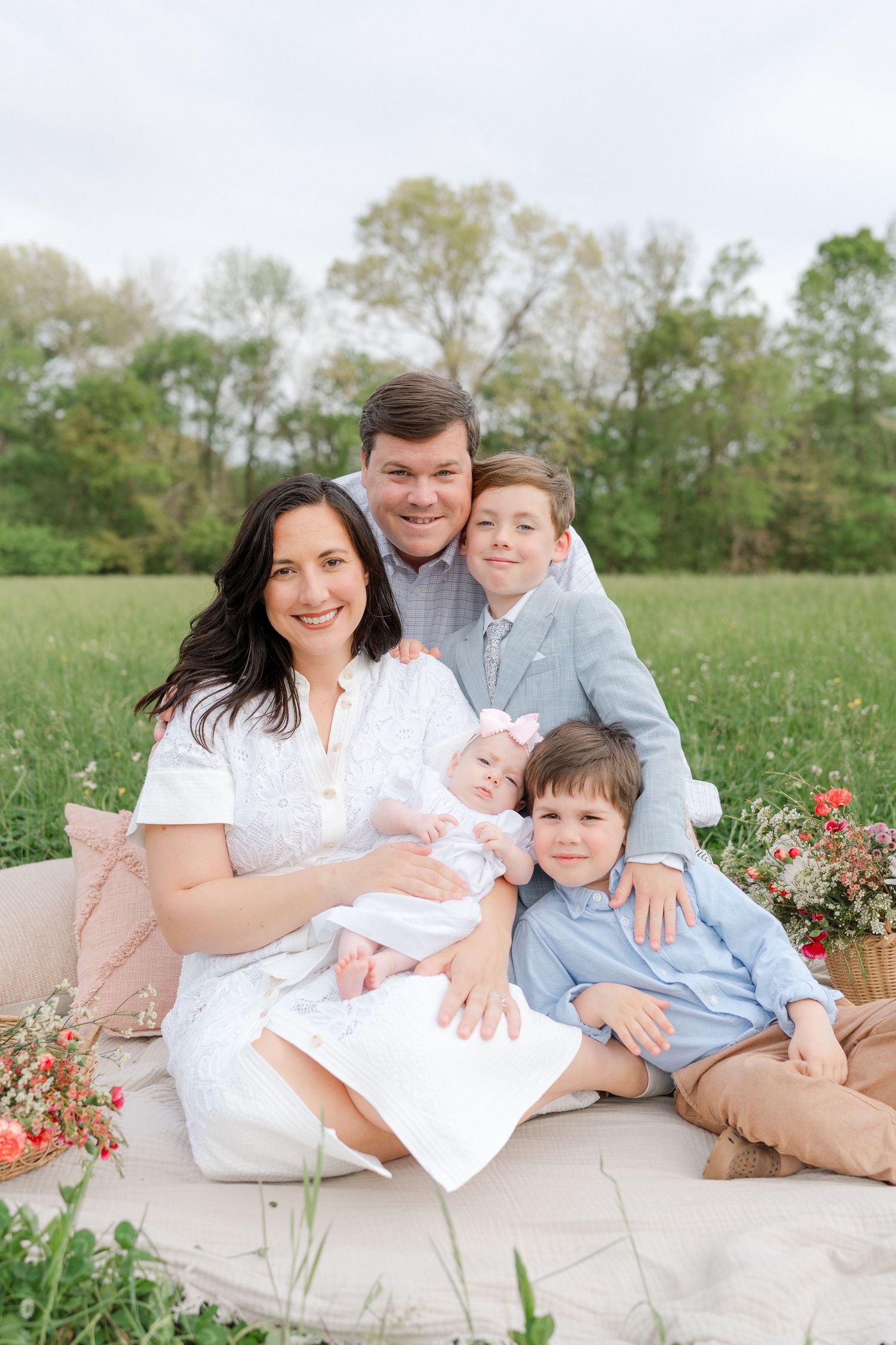 A family of five poses on an outdoor blanket in a green field, with a mother holding a newborn baby.