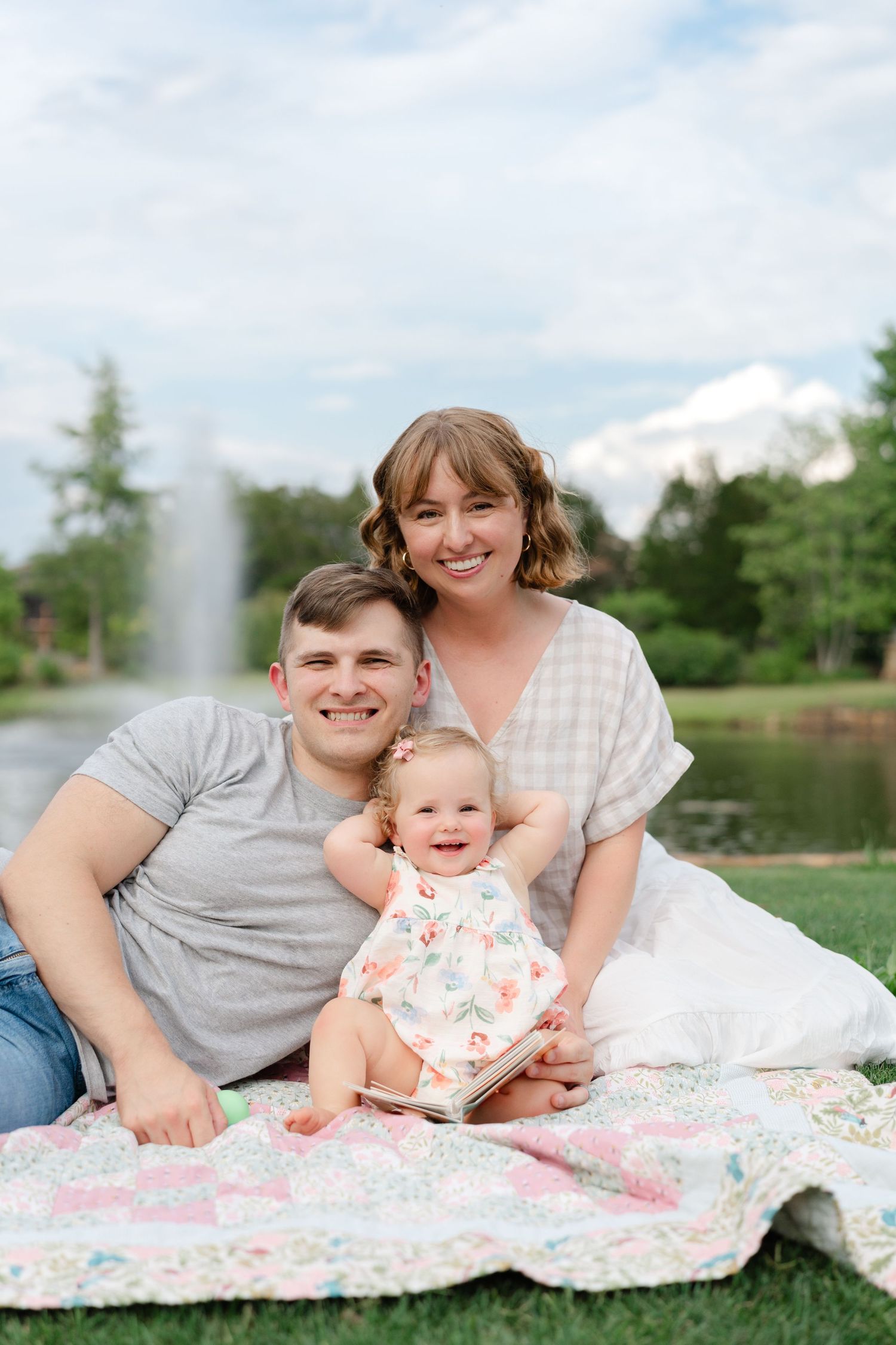 A happy young family with a baby girl sits on a colorful quilt in a park with a fountain backdrop.