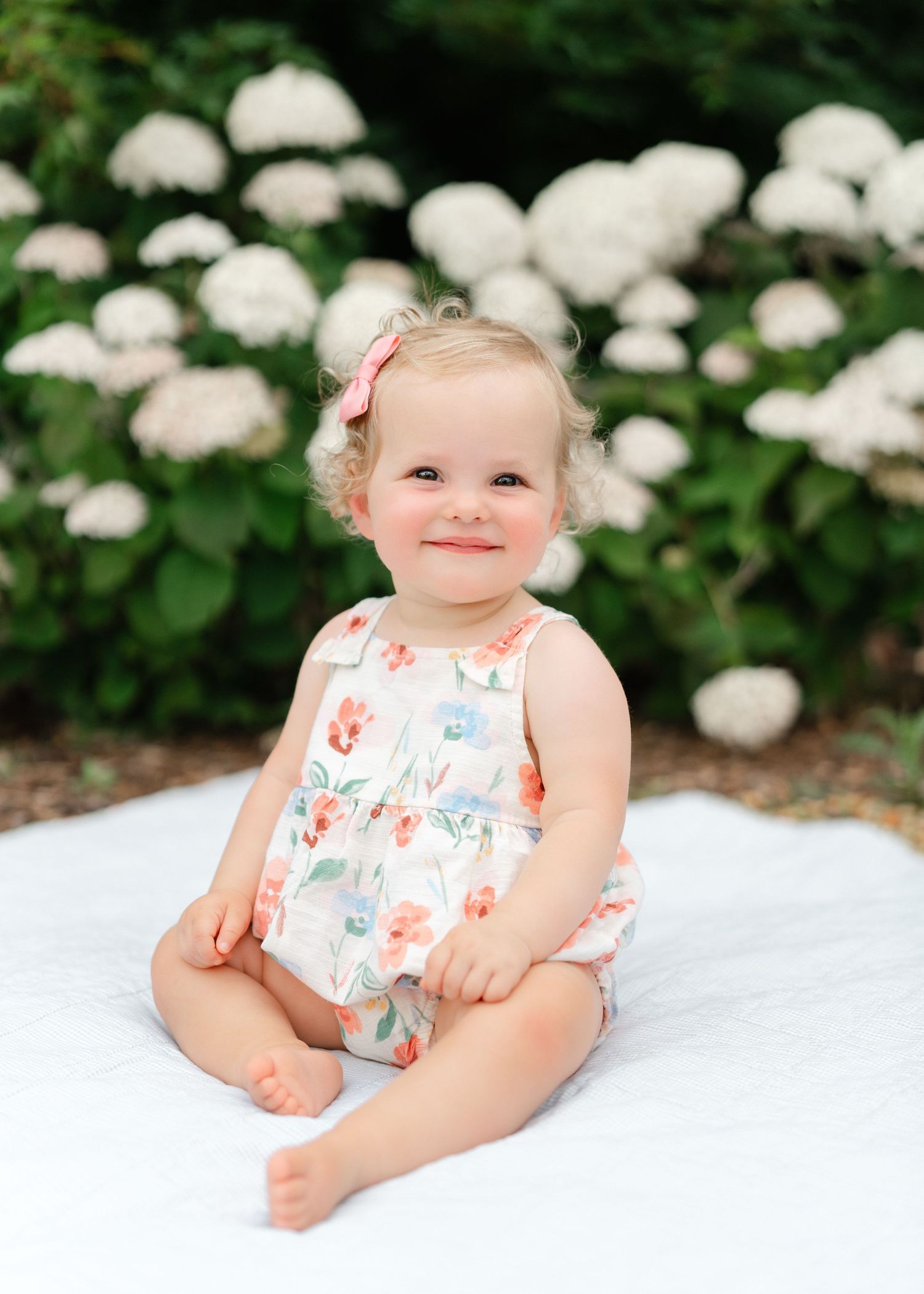 Smiling baby girl in floral romper sitting on white blanket in front of white hydrangea bushes outdoors.