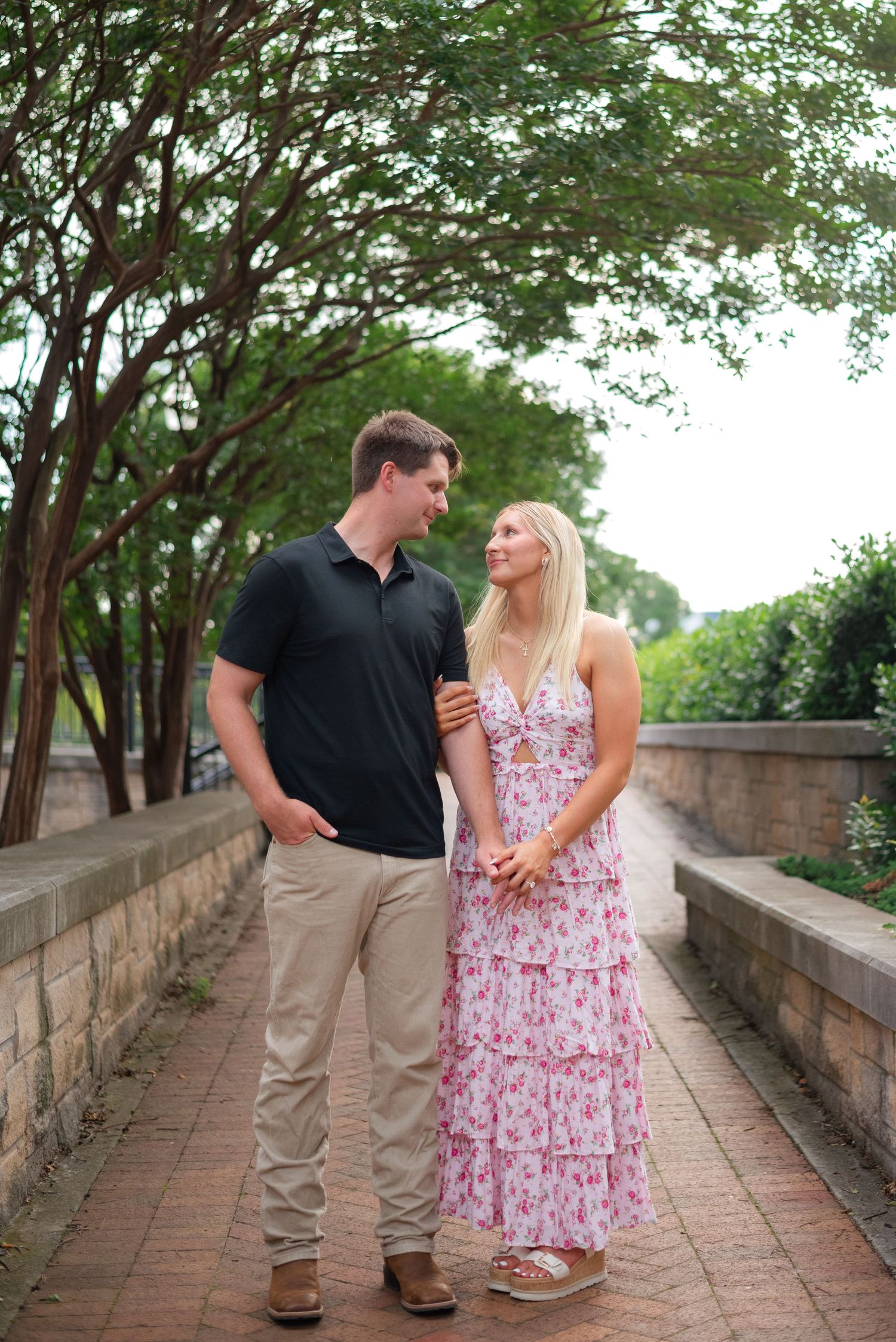 A couple smiles at each other while holding hands on a tree-lined brick path outdoors.