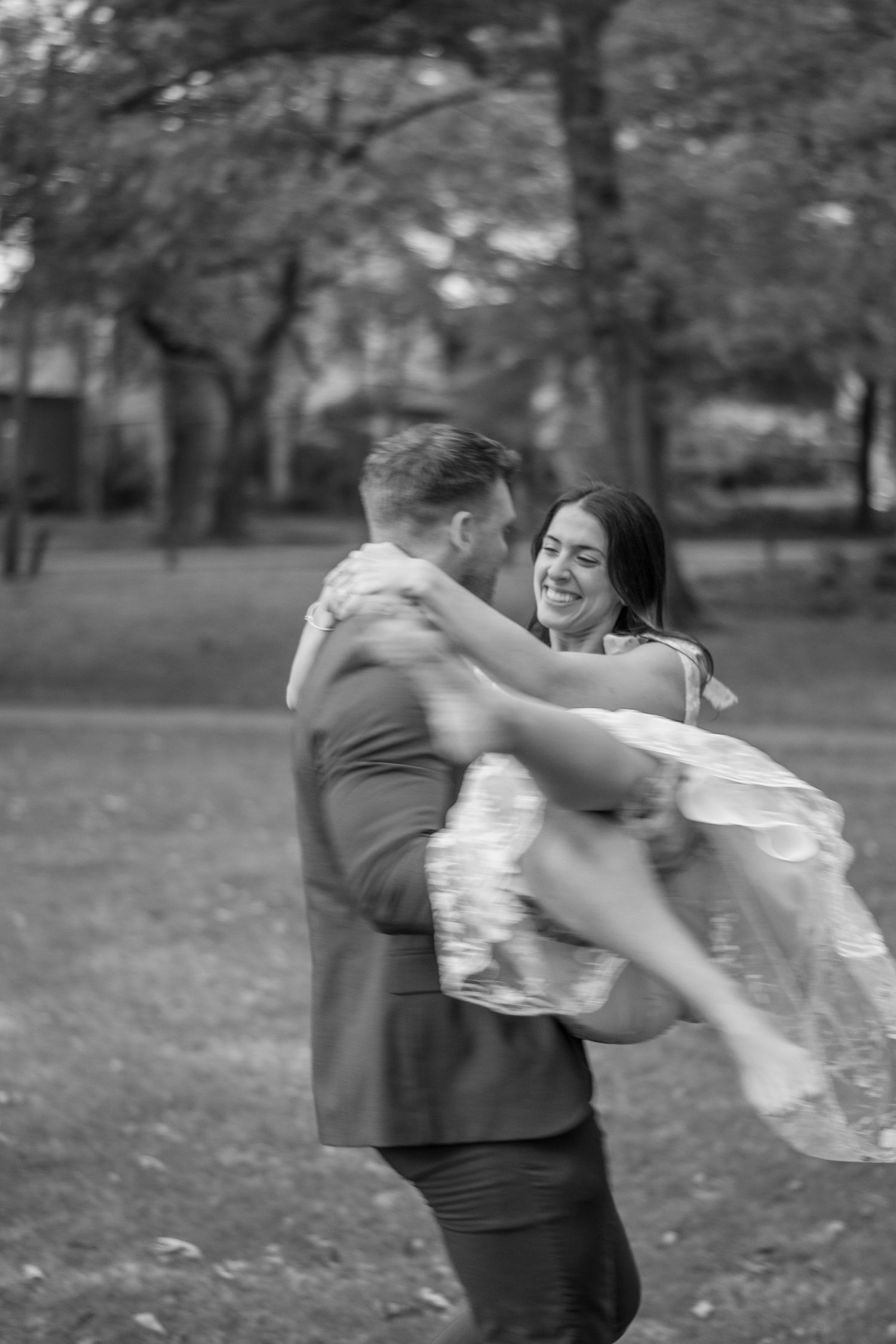 Black and white photo of groom lifting laughing bride in a park, capturing a joyful wedding day moment.