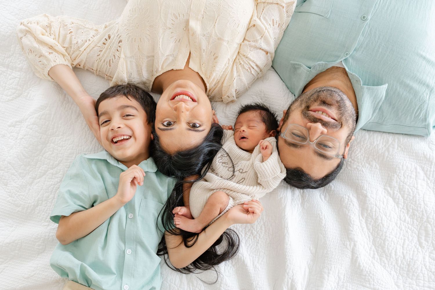 Happy family of four lies together on white bed, parents holding newborn baby with toddler smiling.