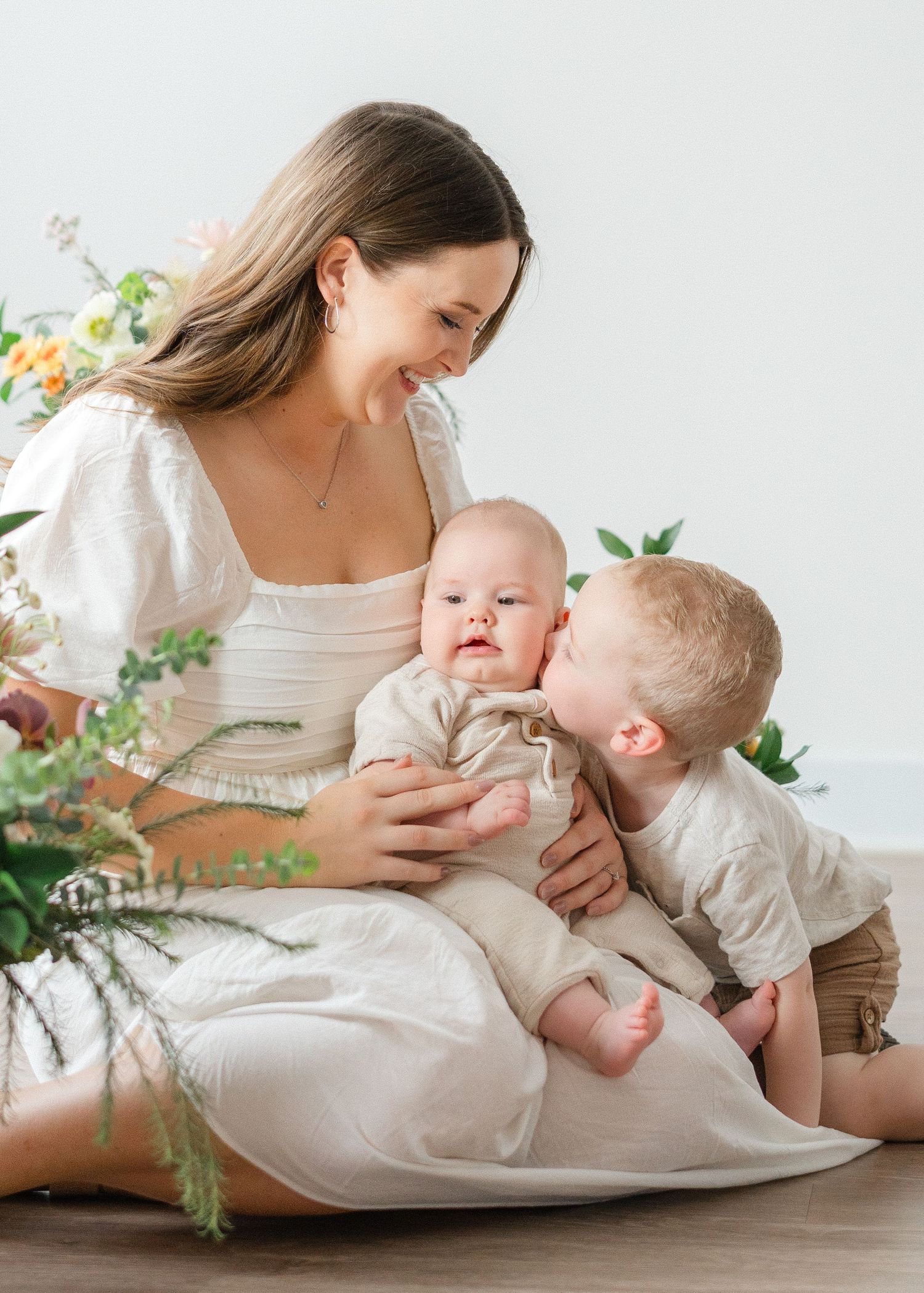 Smiling mother in white dress holds infant while toddler gives baby a kiss beside flowers indoors.
