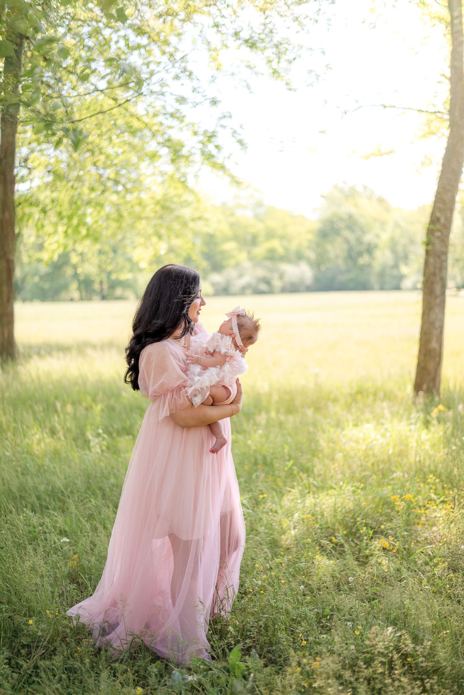 Elegant mother in flowing pink dress cradling newborn baby in a golden sunlit field with tall green trees.