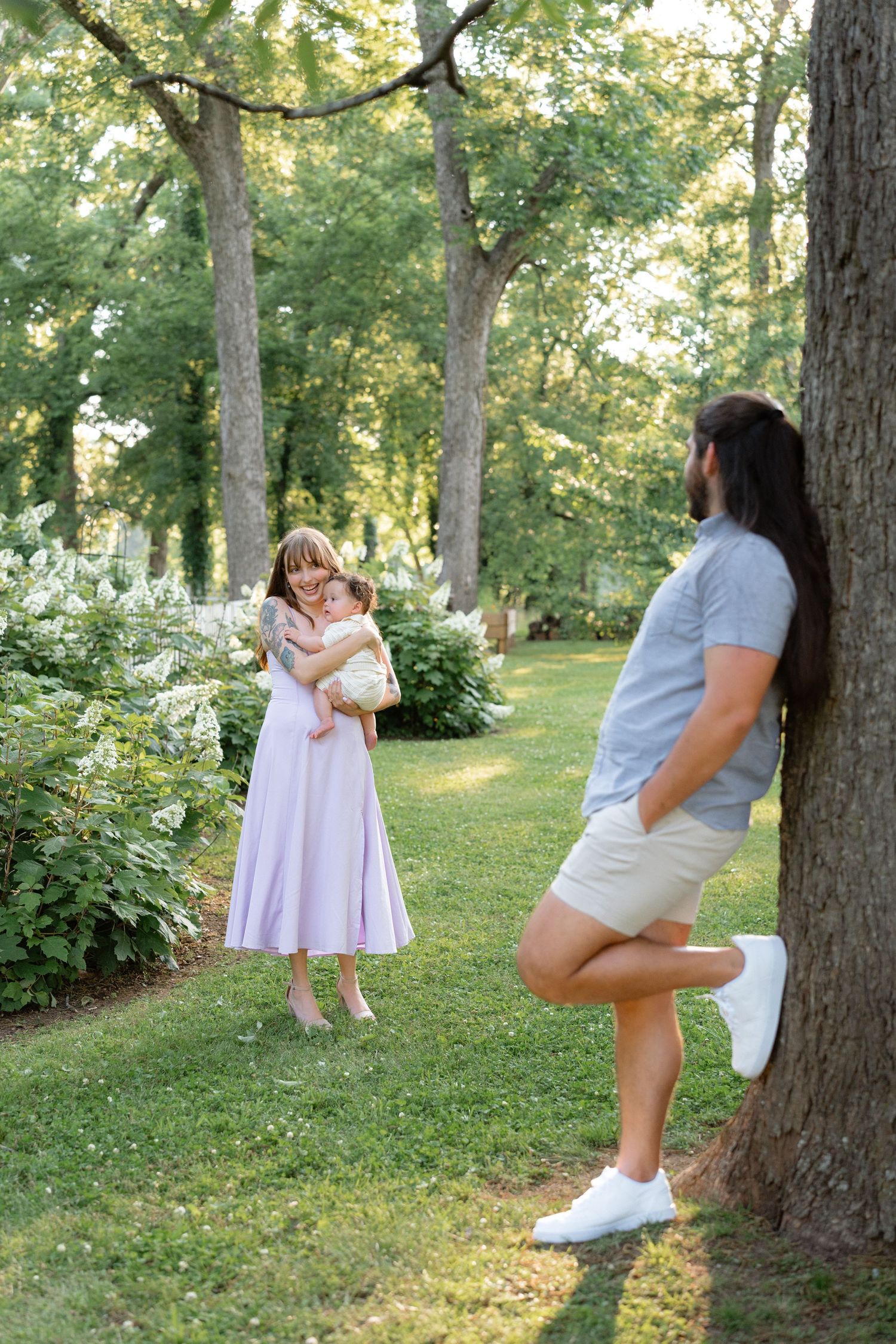 A woman in a lavender dress holds a baby while a bearded man leans against a tree in a lush green park.