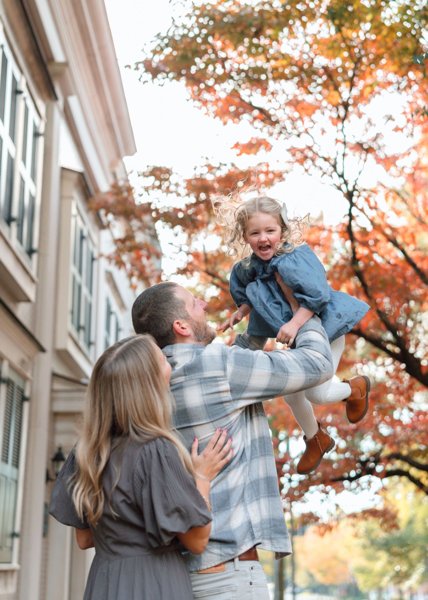 Father lifts laughing daughter in air while mother watches under vibrant fall foliage.