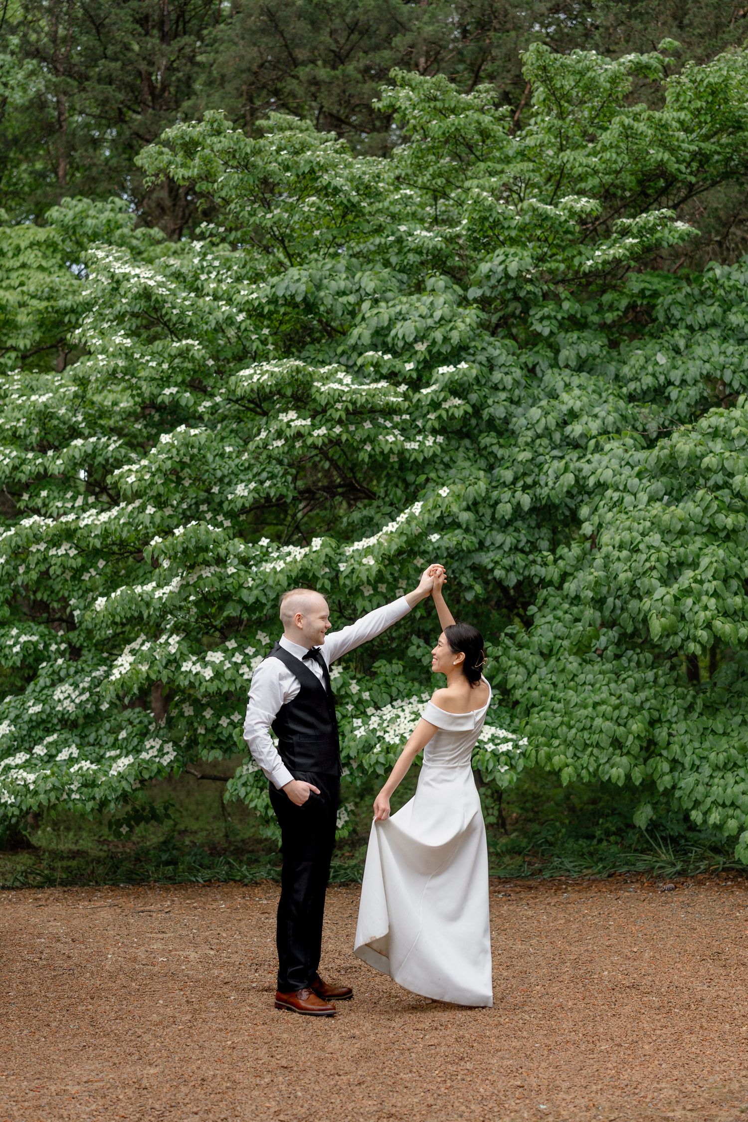 Bride and groom dancing outdoors near blooming white trees on a gravel path in a lush green forest setting.