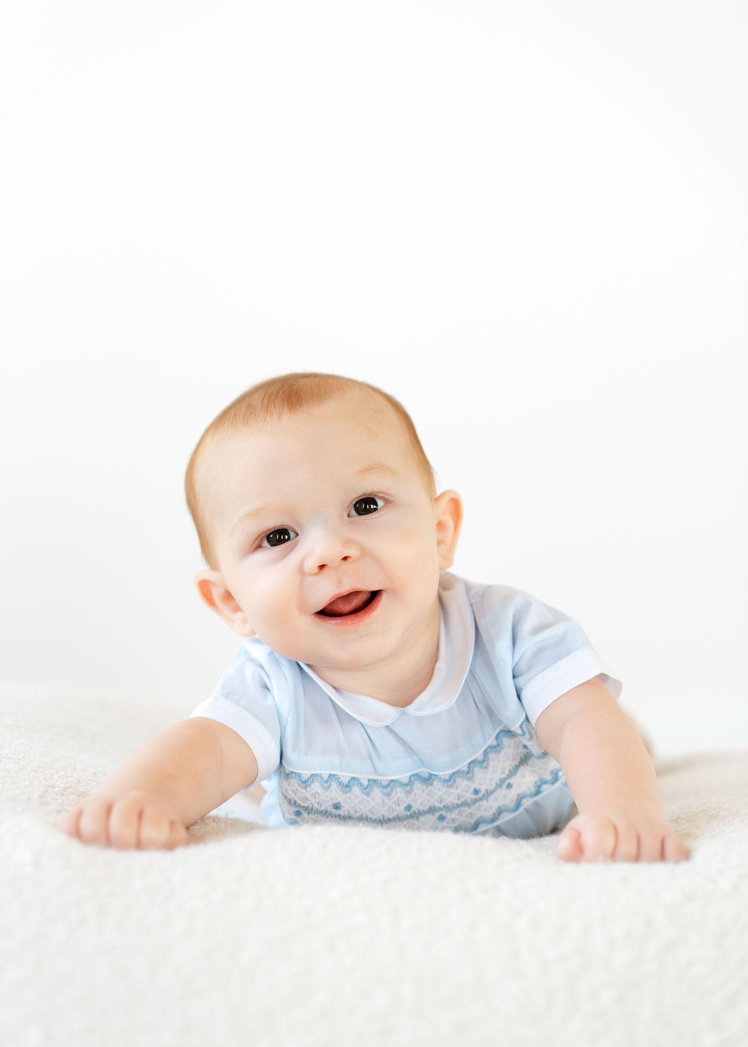 Happy baby boy in a light blue smocked romper doing tummy time on a soft white blanket.