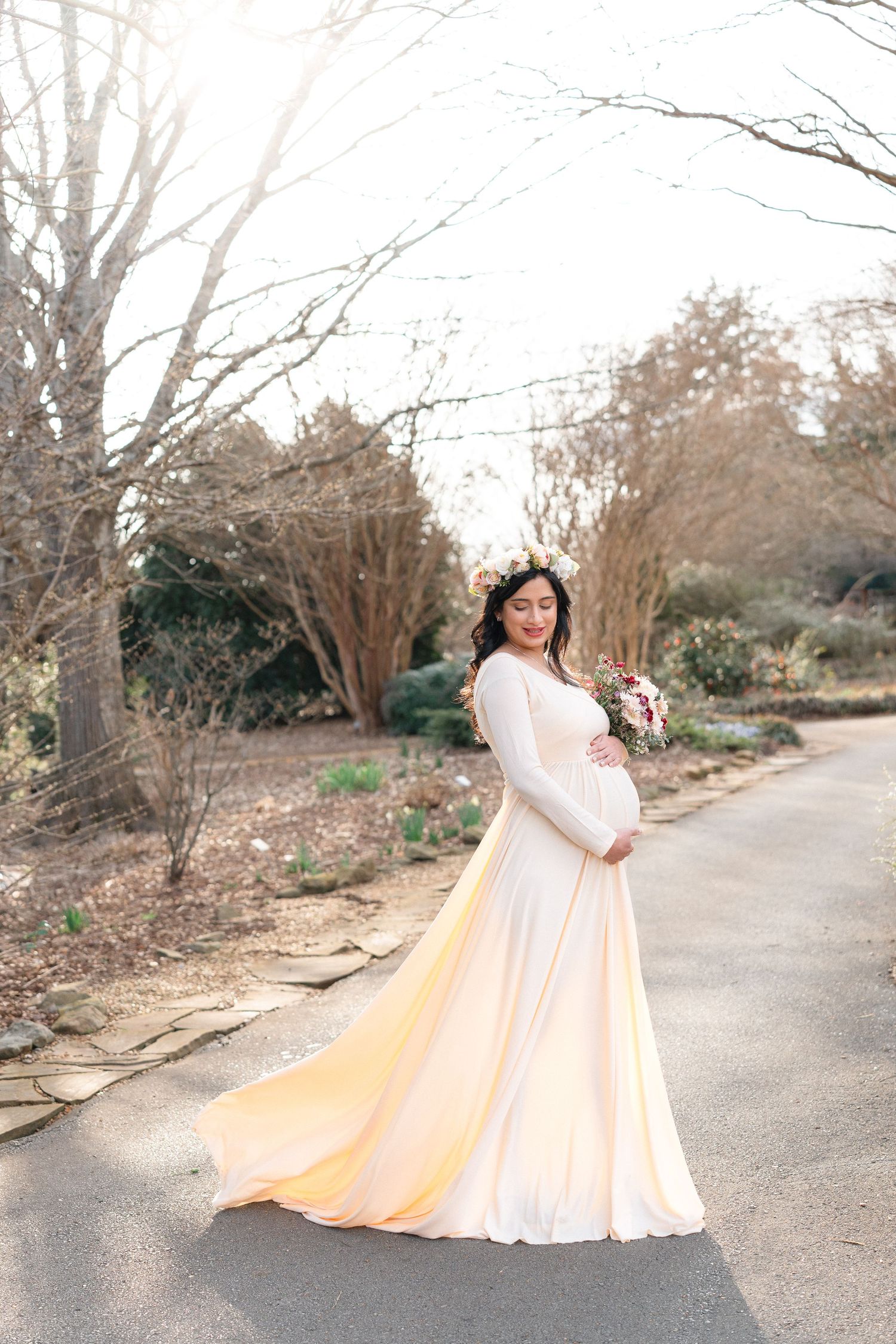 Pregnant woman in flowing cream maternity gown with floral crown poses on garden path.
