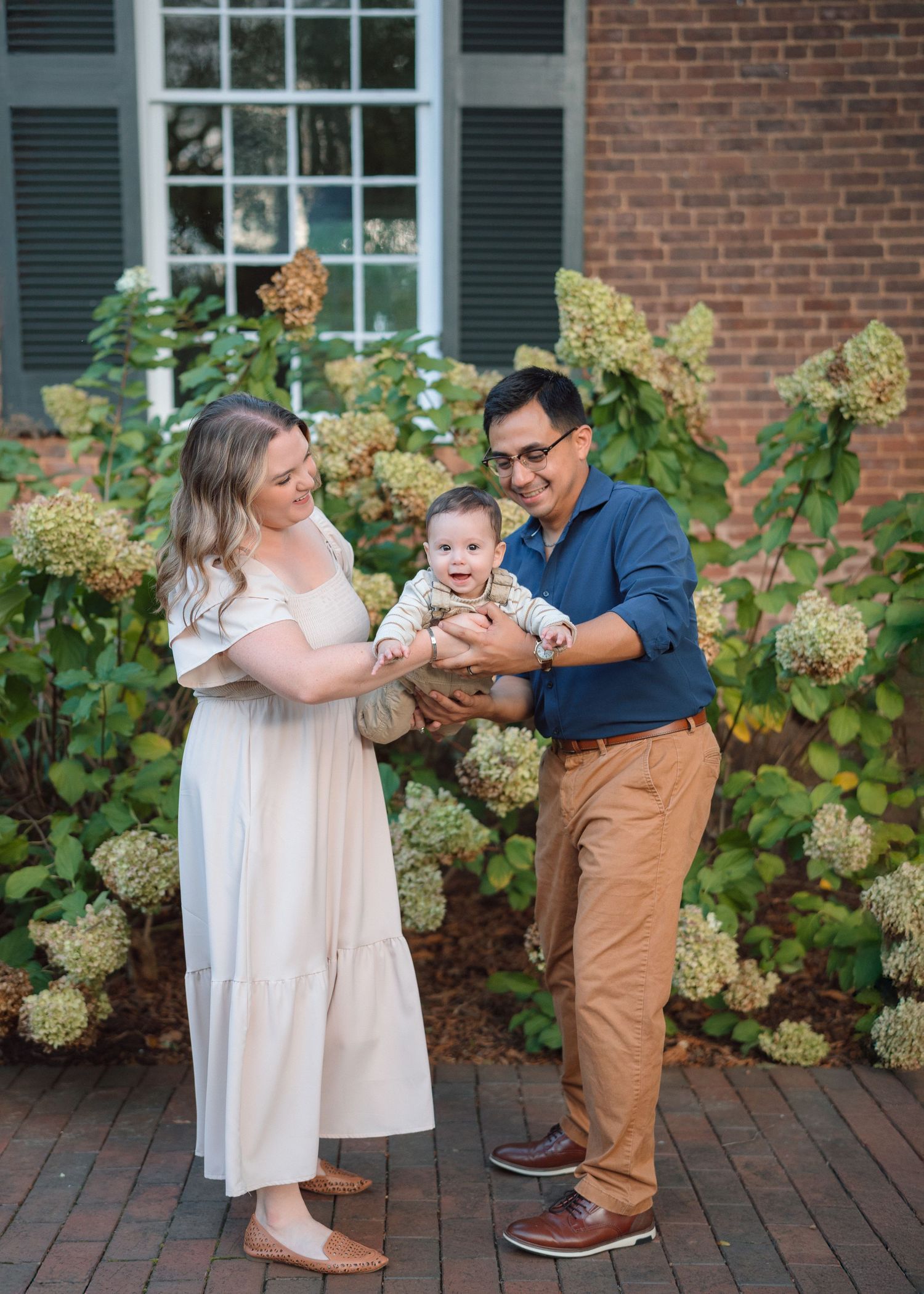 A happy young family with a smiling baby poses outdoors near blooming hydrangeas and a brick building.