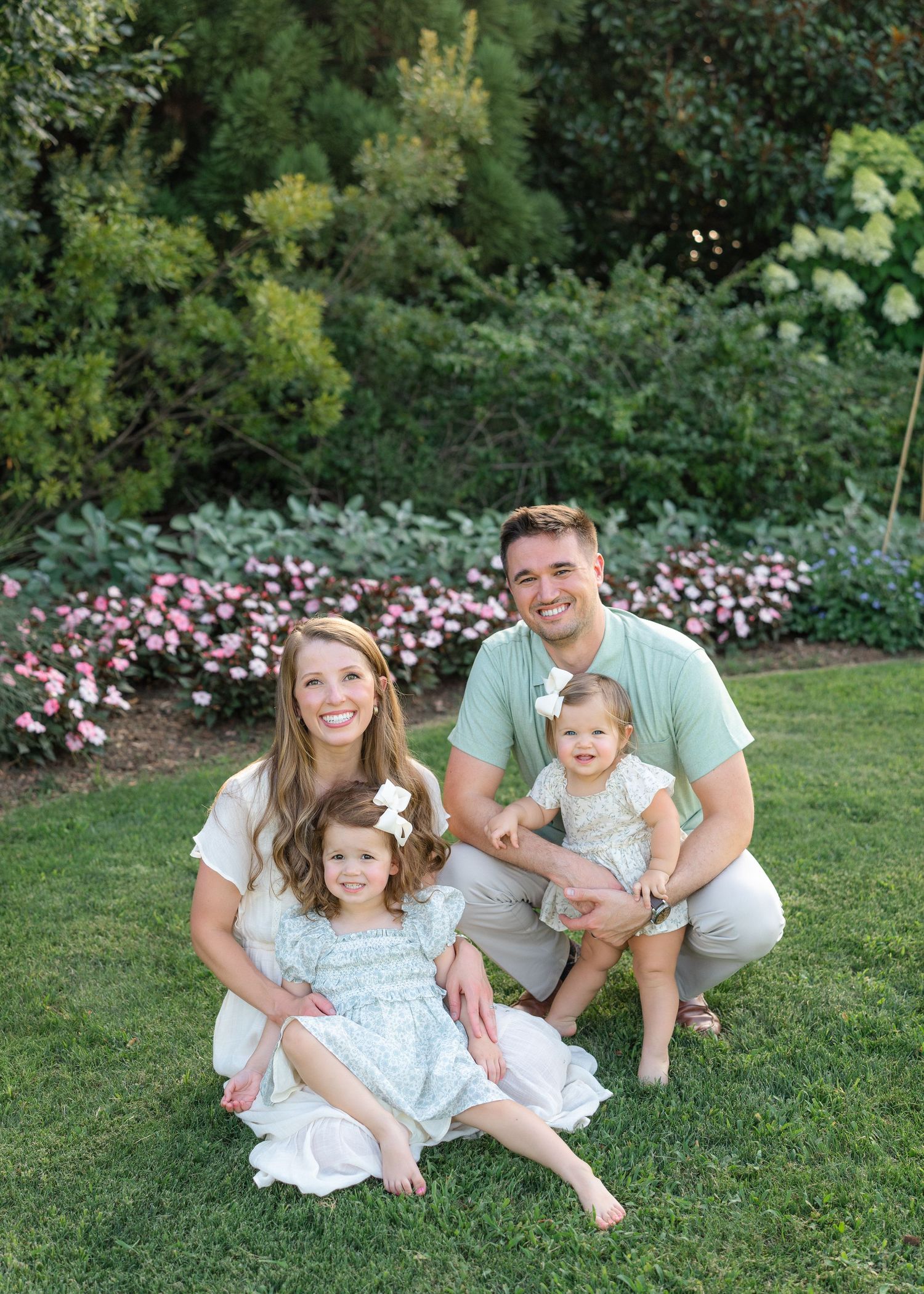 A smiling family of four poses on green grass in front of a lush garden with pink flowers in bloom.
