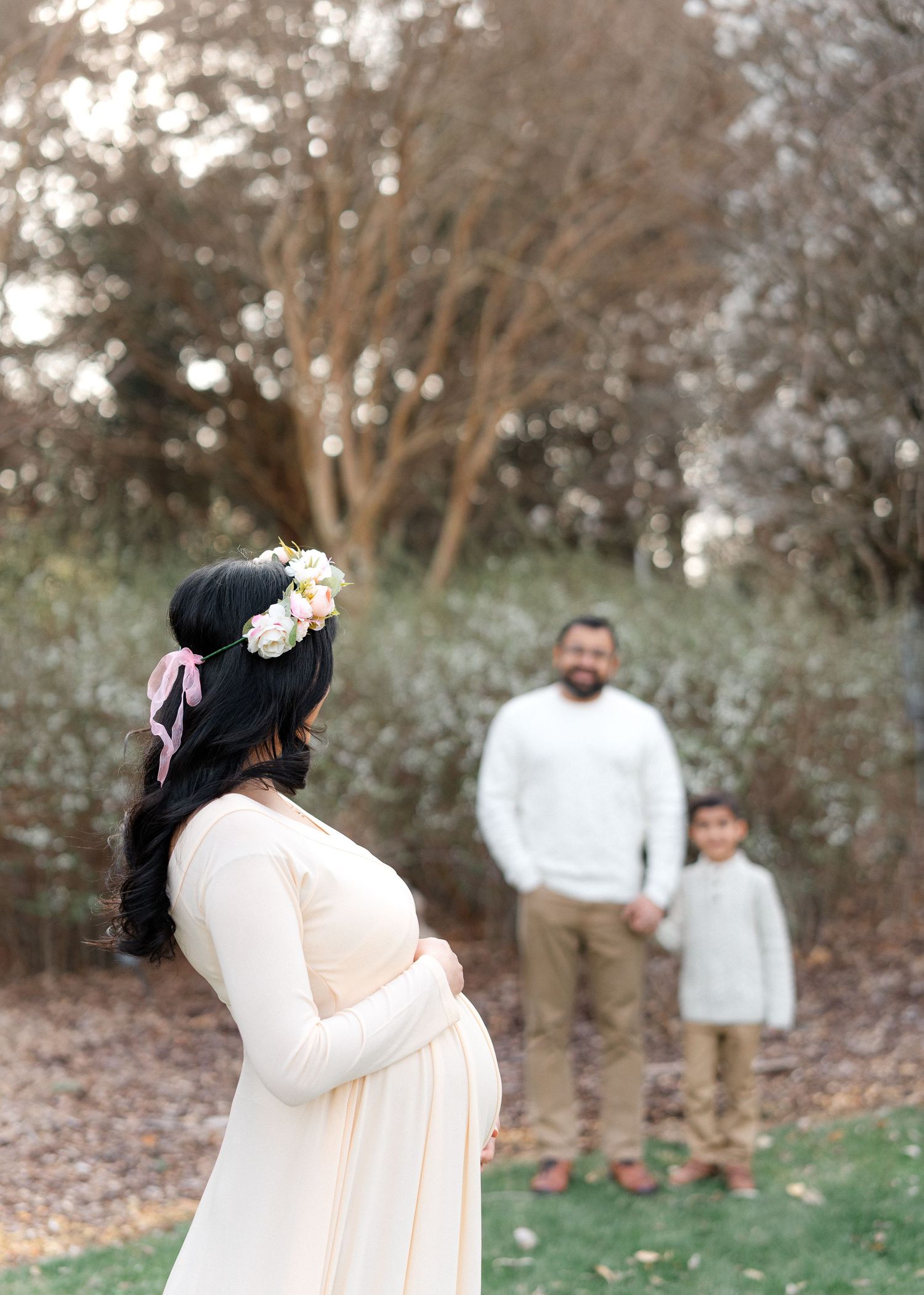 Pregnant woman in cream dress with floral crown faces husband and son in garden.