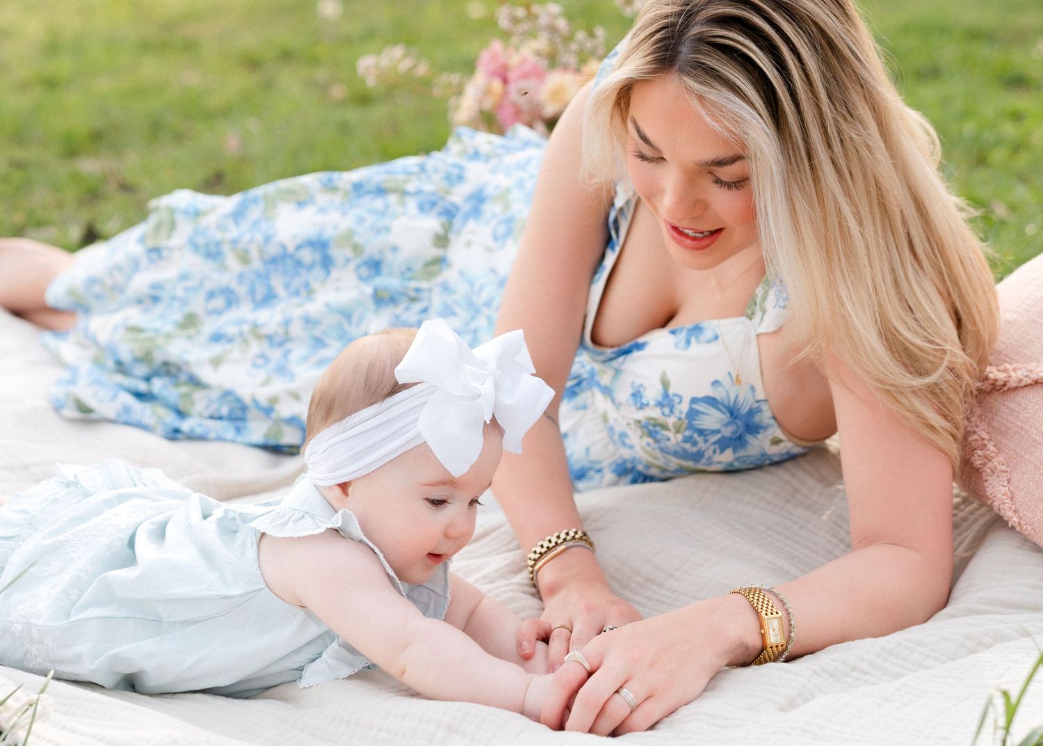 Mother in blue floral dress lies on blanket smiling at baby girl with white bow headband.