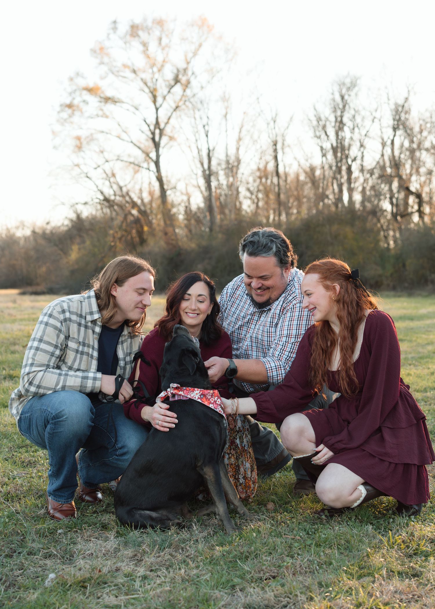 A family of four crouches together petting their black dog in an open field at sunset.